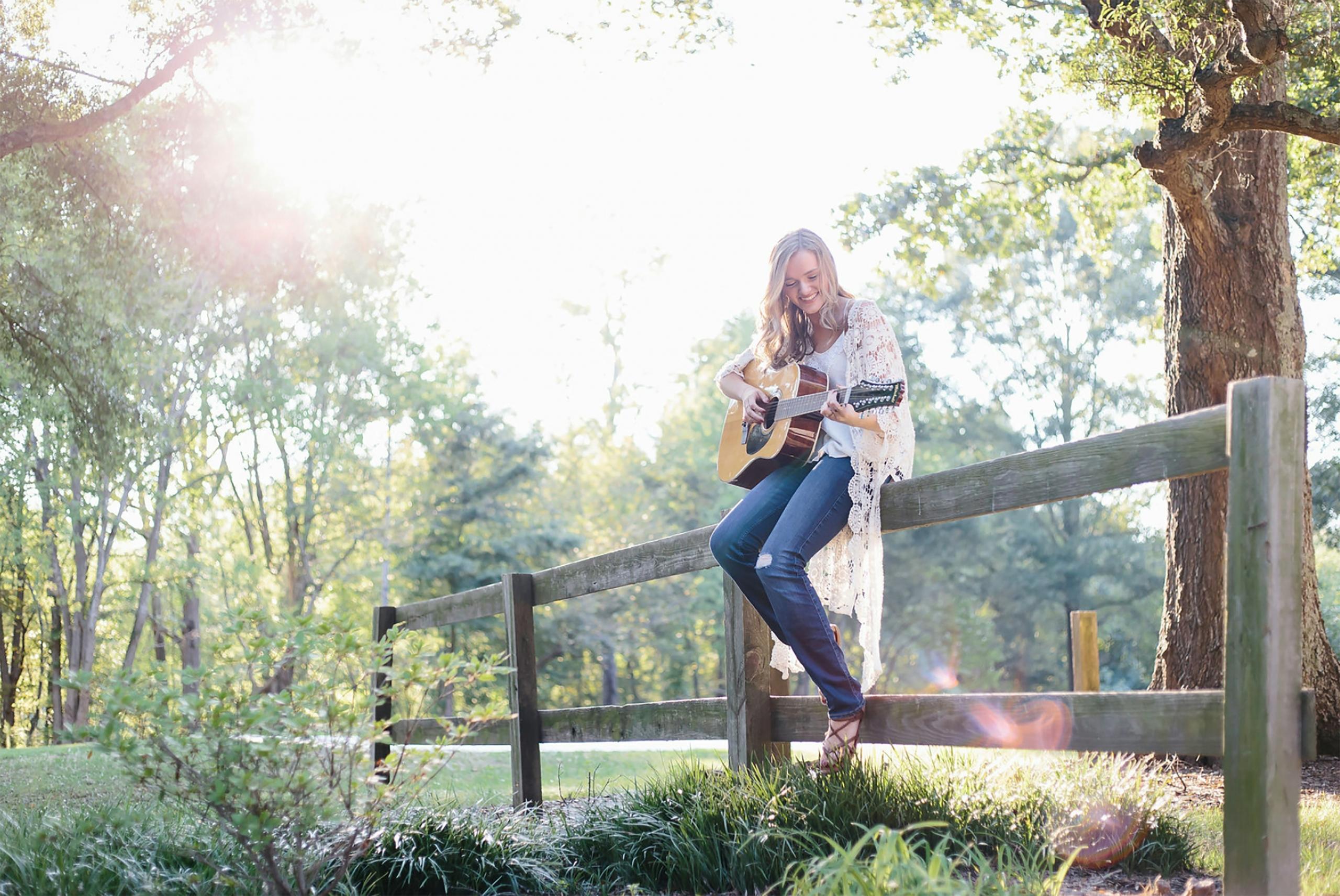 En dame som sitter på et gjerde i naturen og spiller gitar.