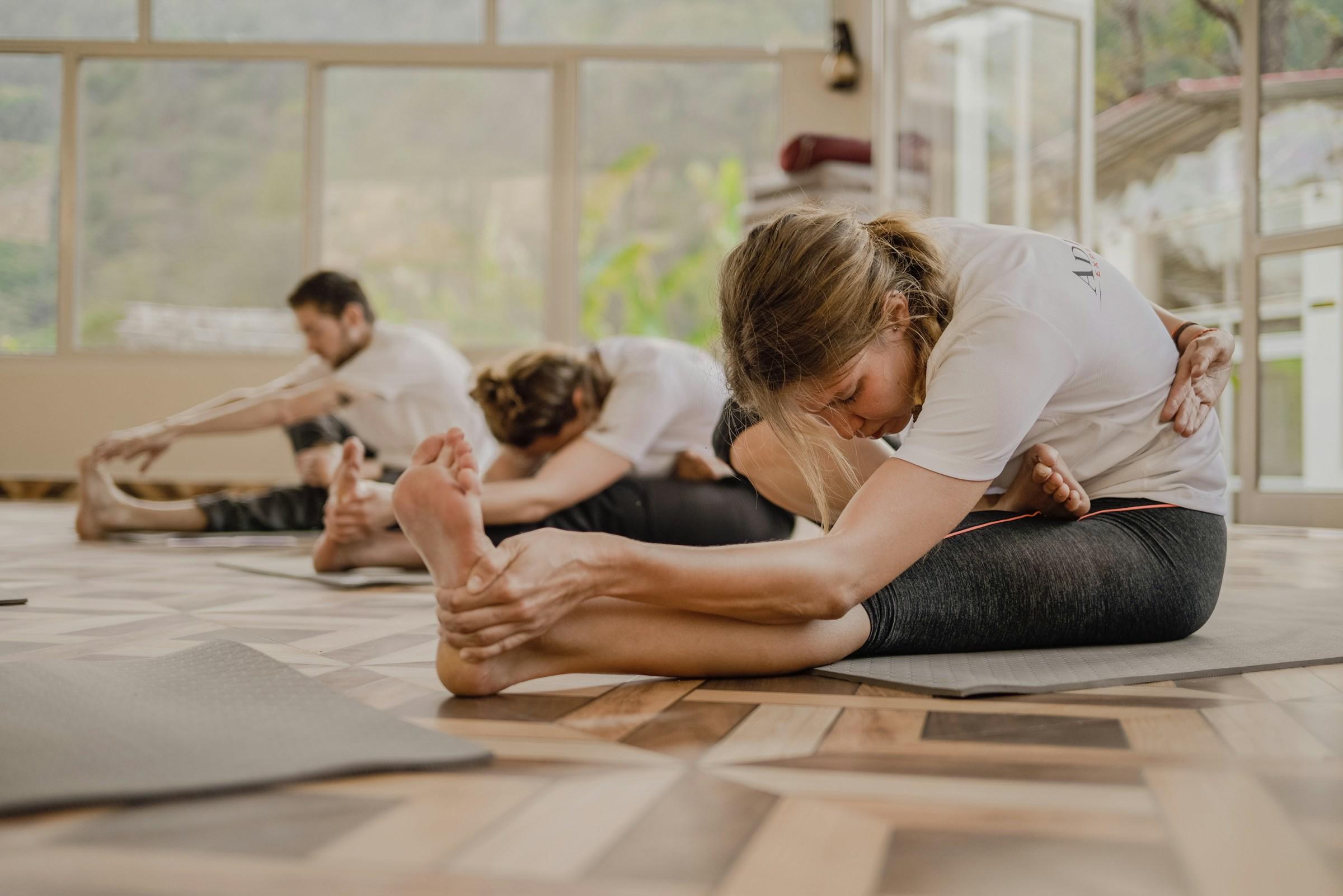Tre personer i et yogastudio som sitter på gulvet og tøyer. 
