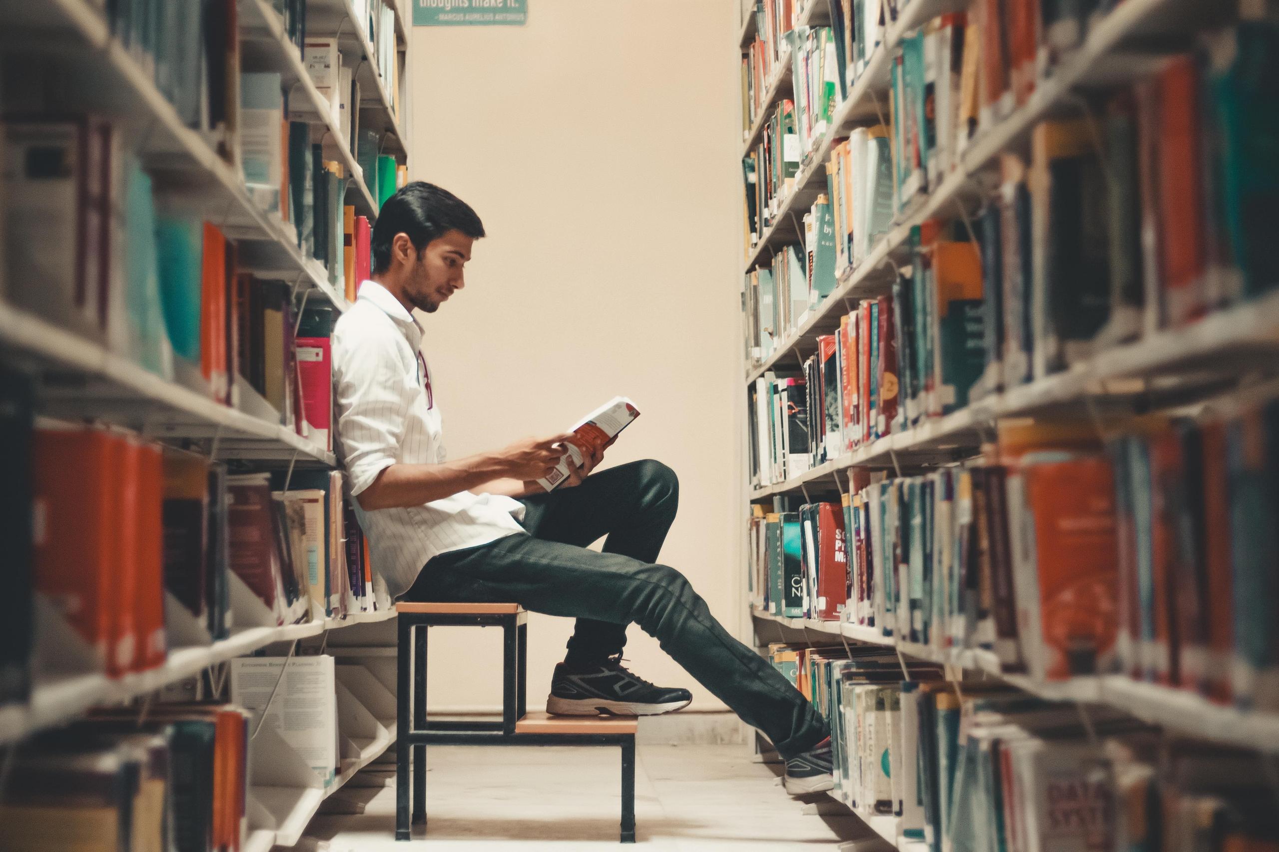 en mann sitter mellom hylleradene i et bibliotek og leser i en bok