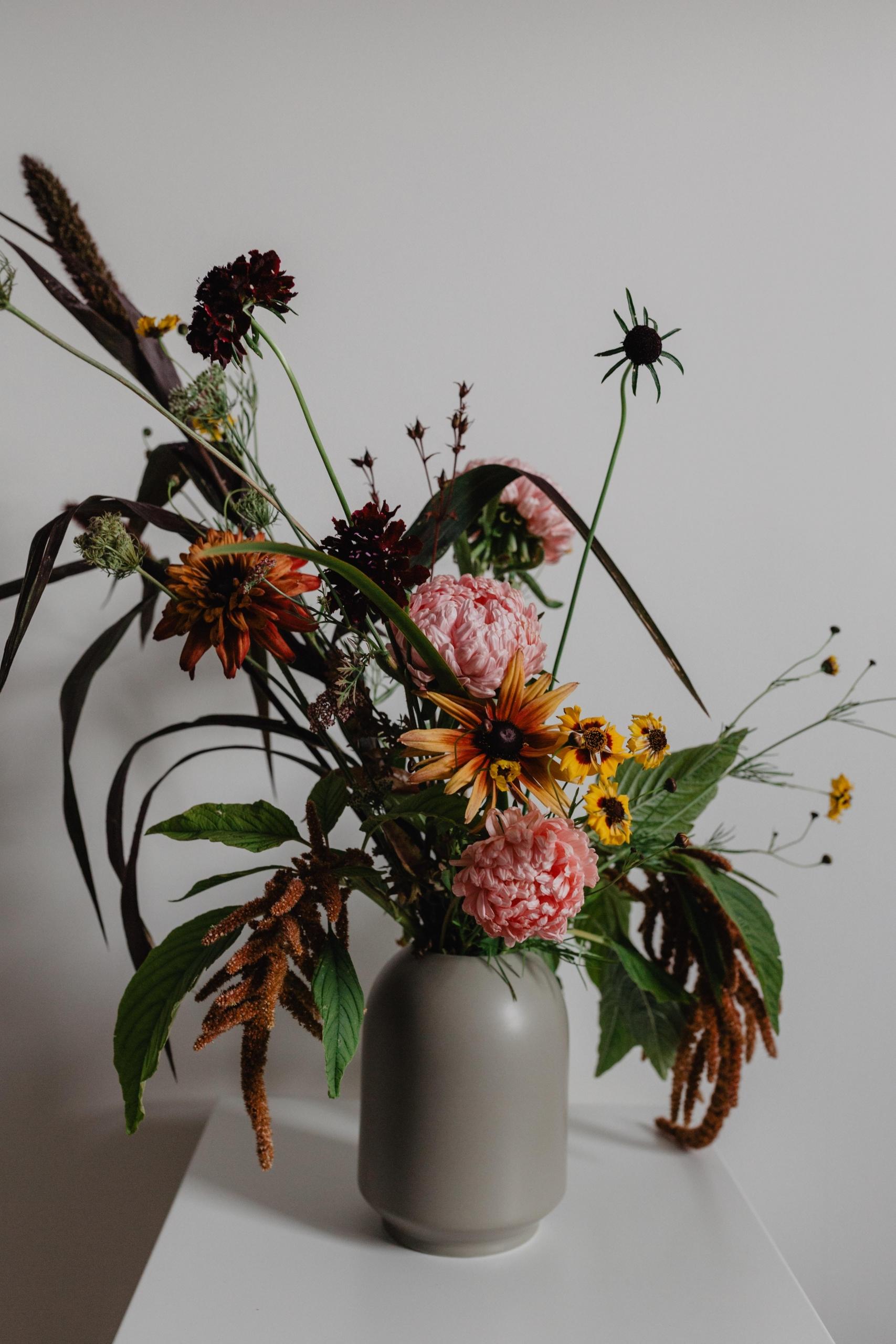en grå vase med høye blomster står på et bord, grønt, gult og rosa