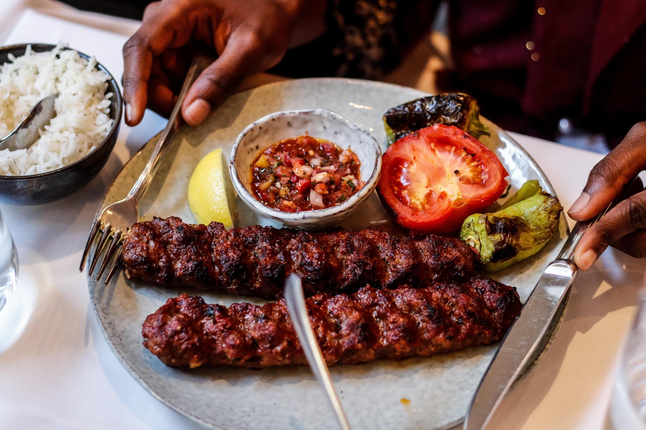 En tallerken med to store kjøttstykker, en halv tomat og flere grillede grønnsaker, ved siden av står en skål med ris. Et par hender er klare til å spise