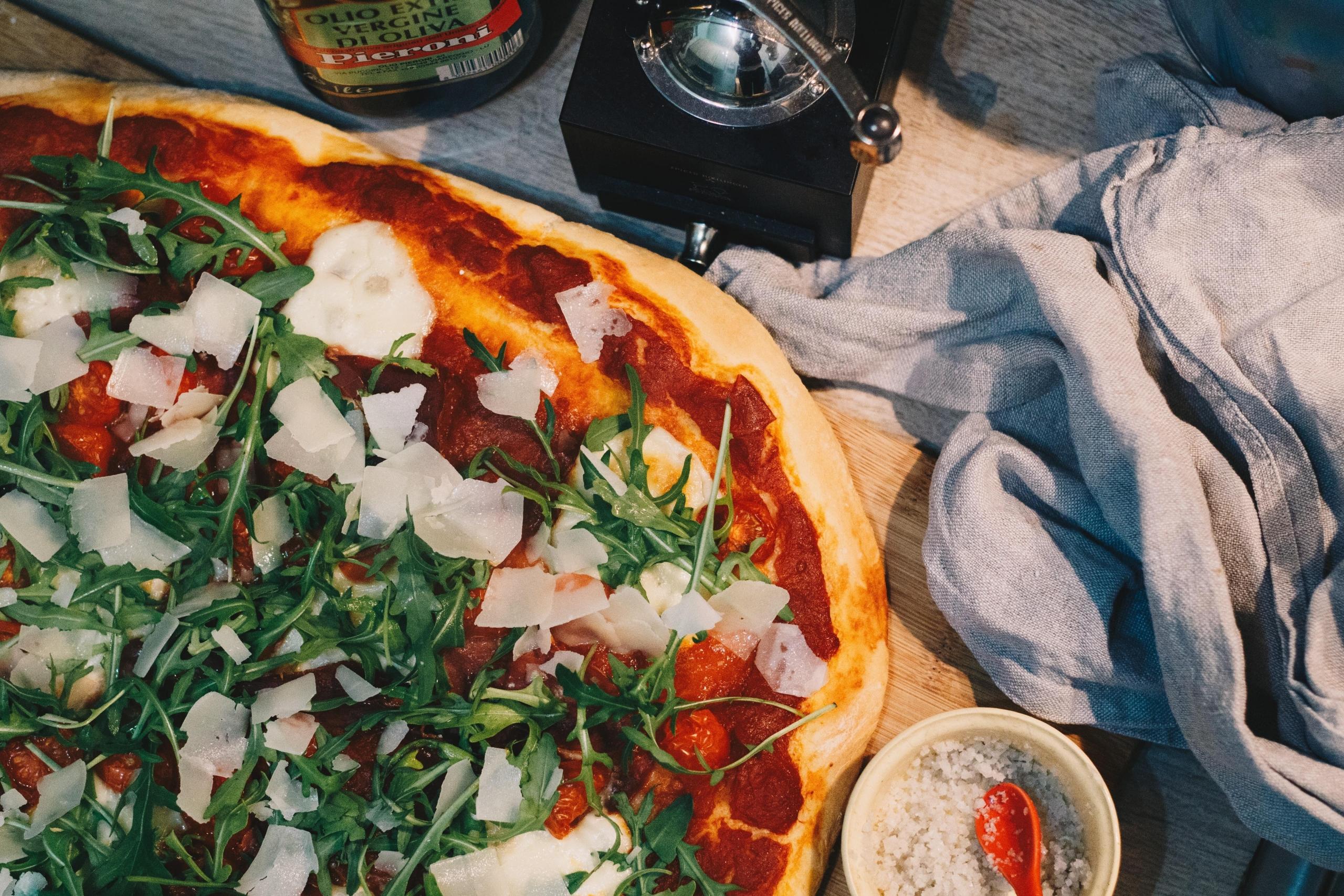 bilde ovenfra av en pizza med tomatsaus, ruccola, mozzarella og parmesan, en flaske olivenolje står i toppen av bildet