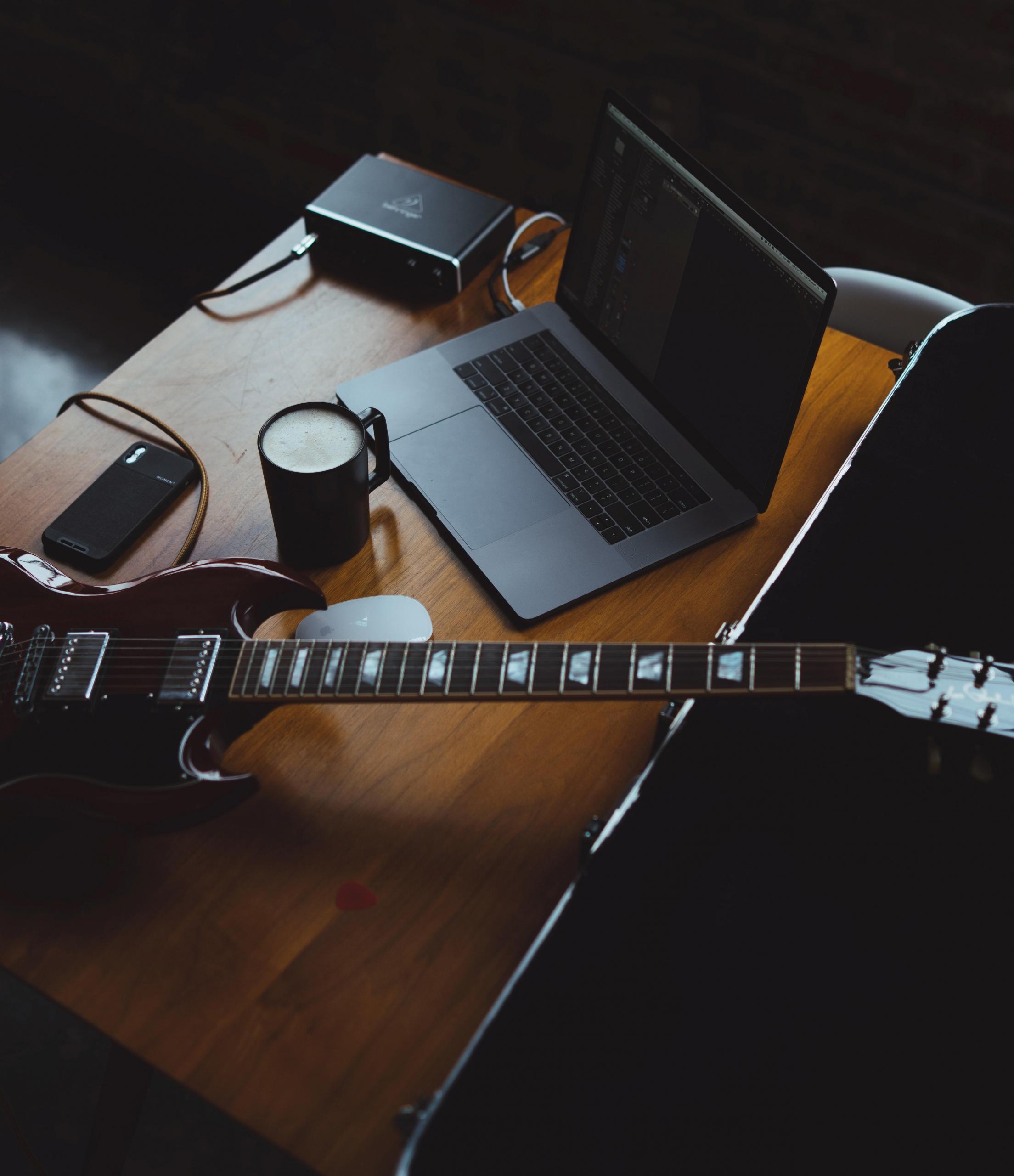 en elektrisk gitar ligger på et bord sammen med en laptop, en kopp med varm drikke og en mobiltelefon