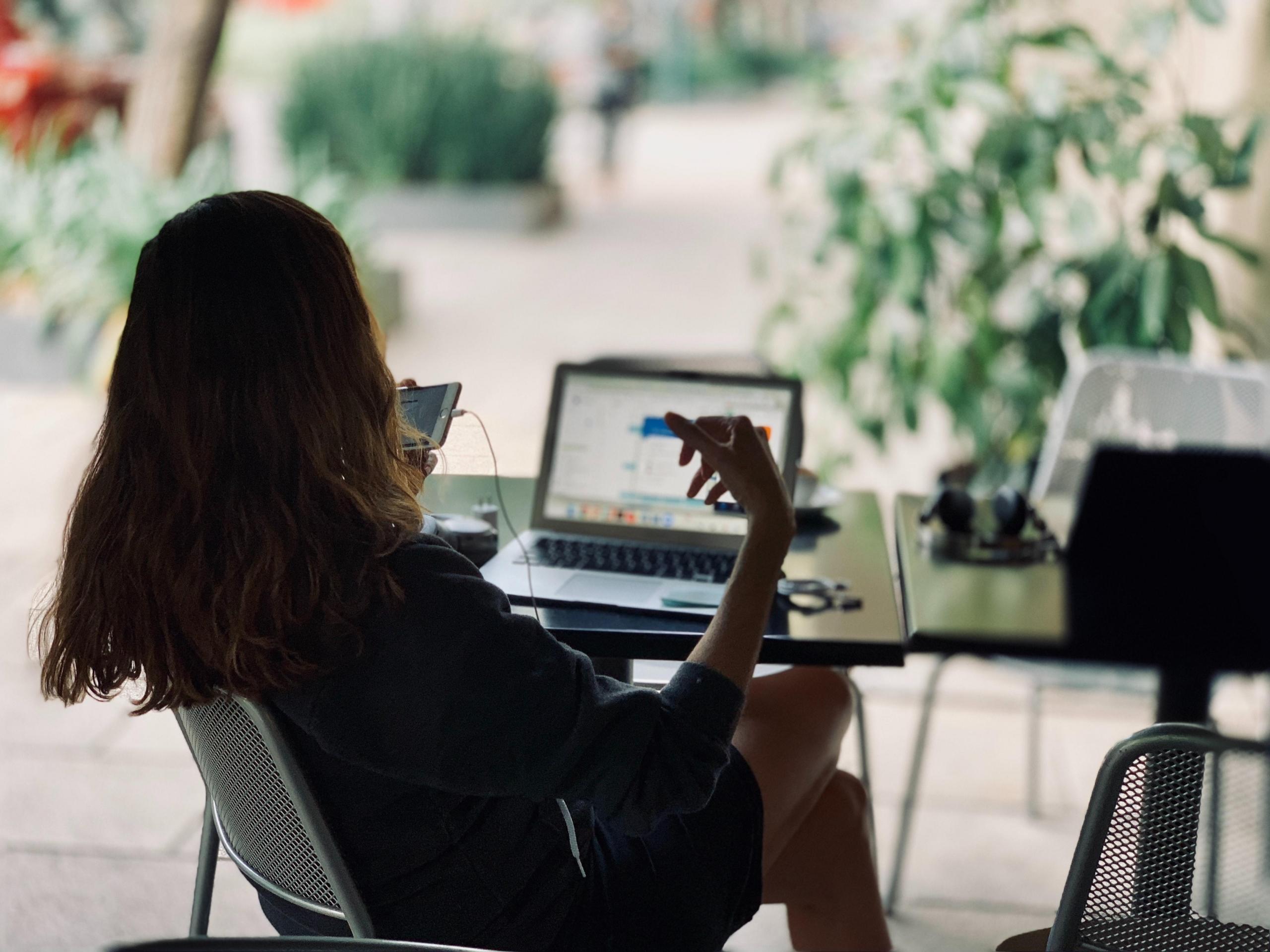 Kvinne sitter ved et kafébord med en mobil foran ansiktet, på bordet står en åpen laptop, hun interagerer med begge to
