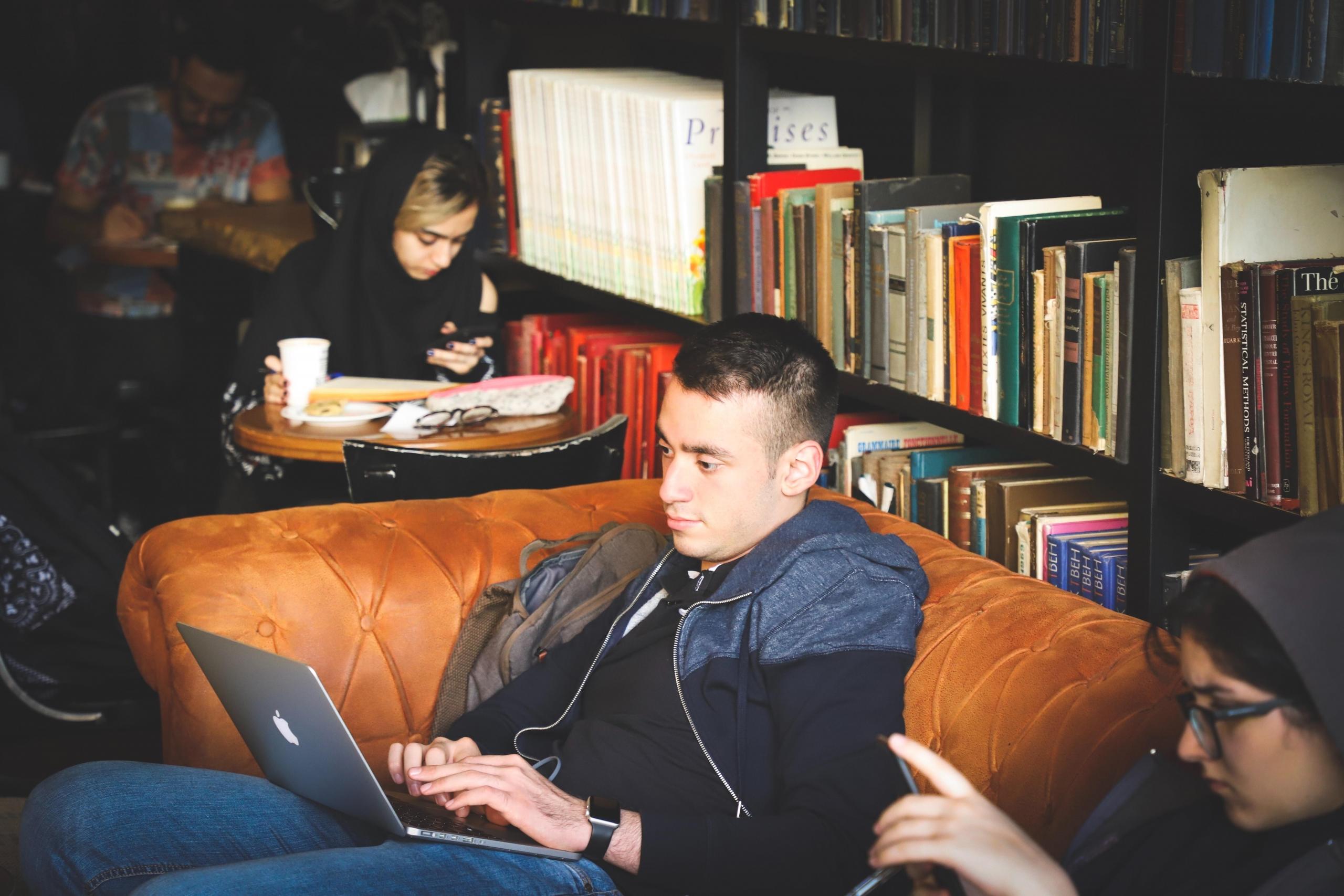 mann sitter med pc på fanget i en sofa på en kafé eller i et bibliotek, flere konsentrerte mennesker sitter i nærheten, bak sofaen står en bred bokhylle