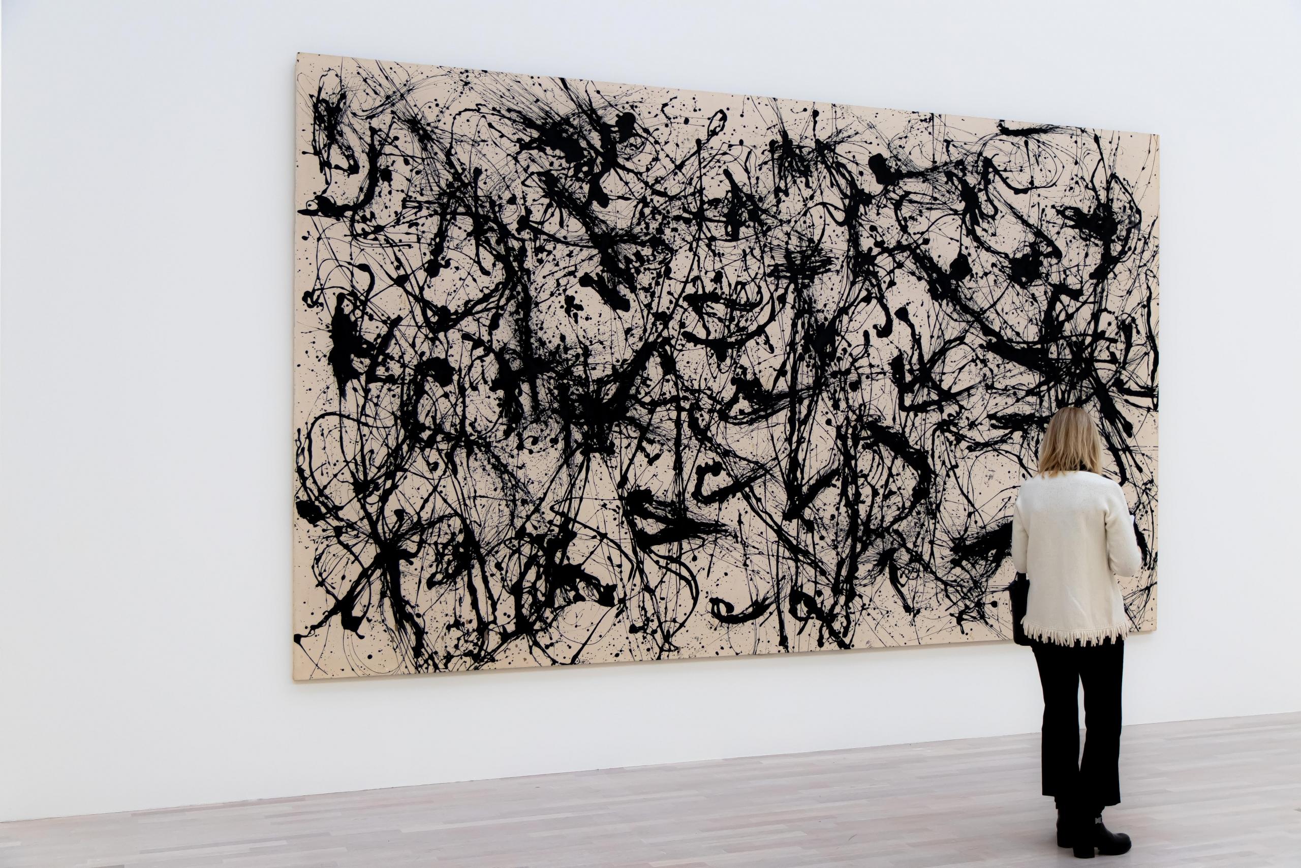 et stort hvitt og svart maleri henger på en vegg i et galleri, en kvinne står og ser på det