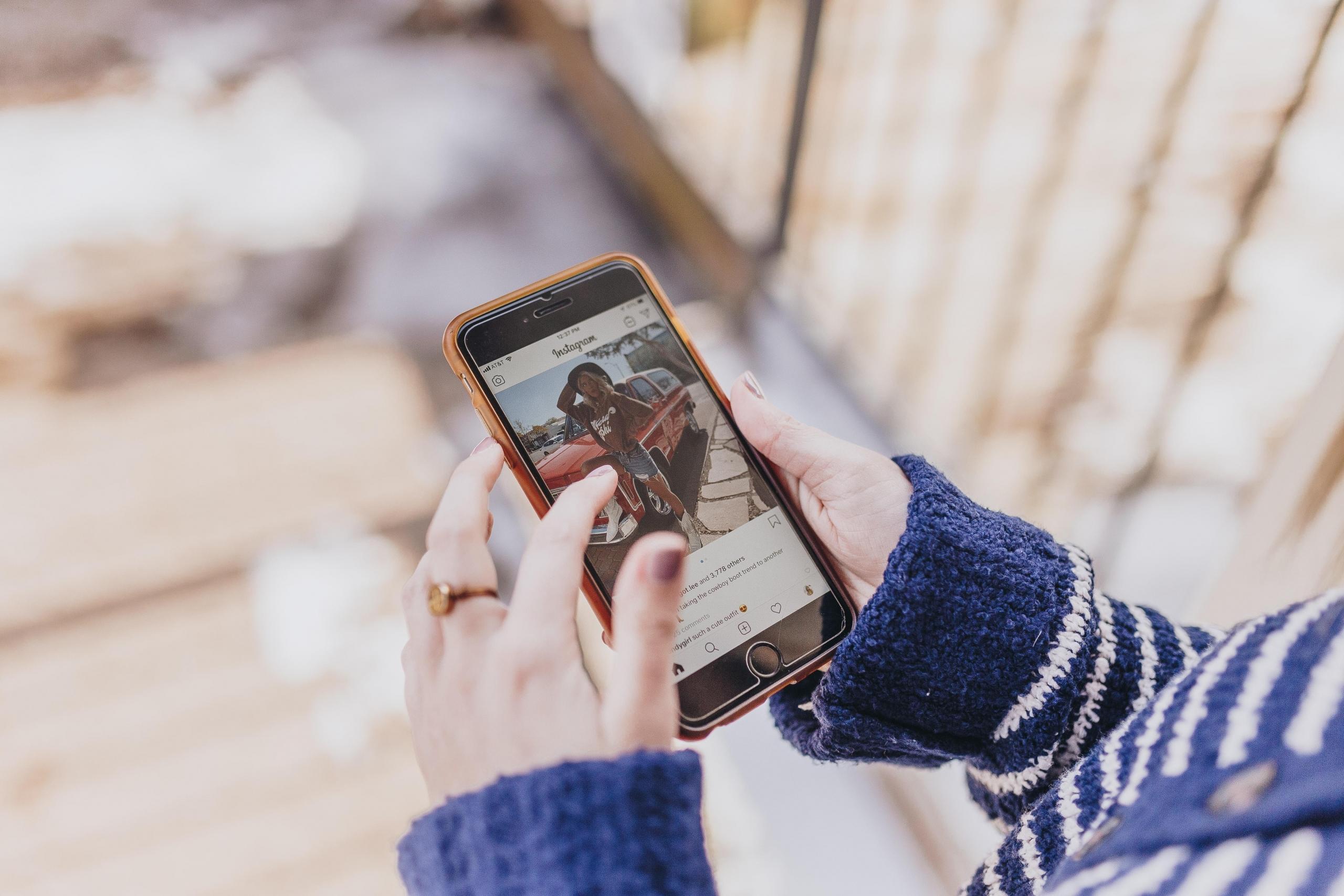 To hender holder i en mobil som hår Instagram-appen åpen. Skjermen viser et Instagram-bilde av en dame i shorts.