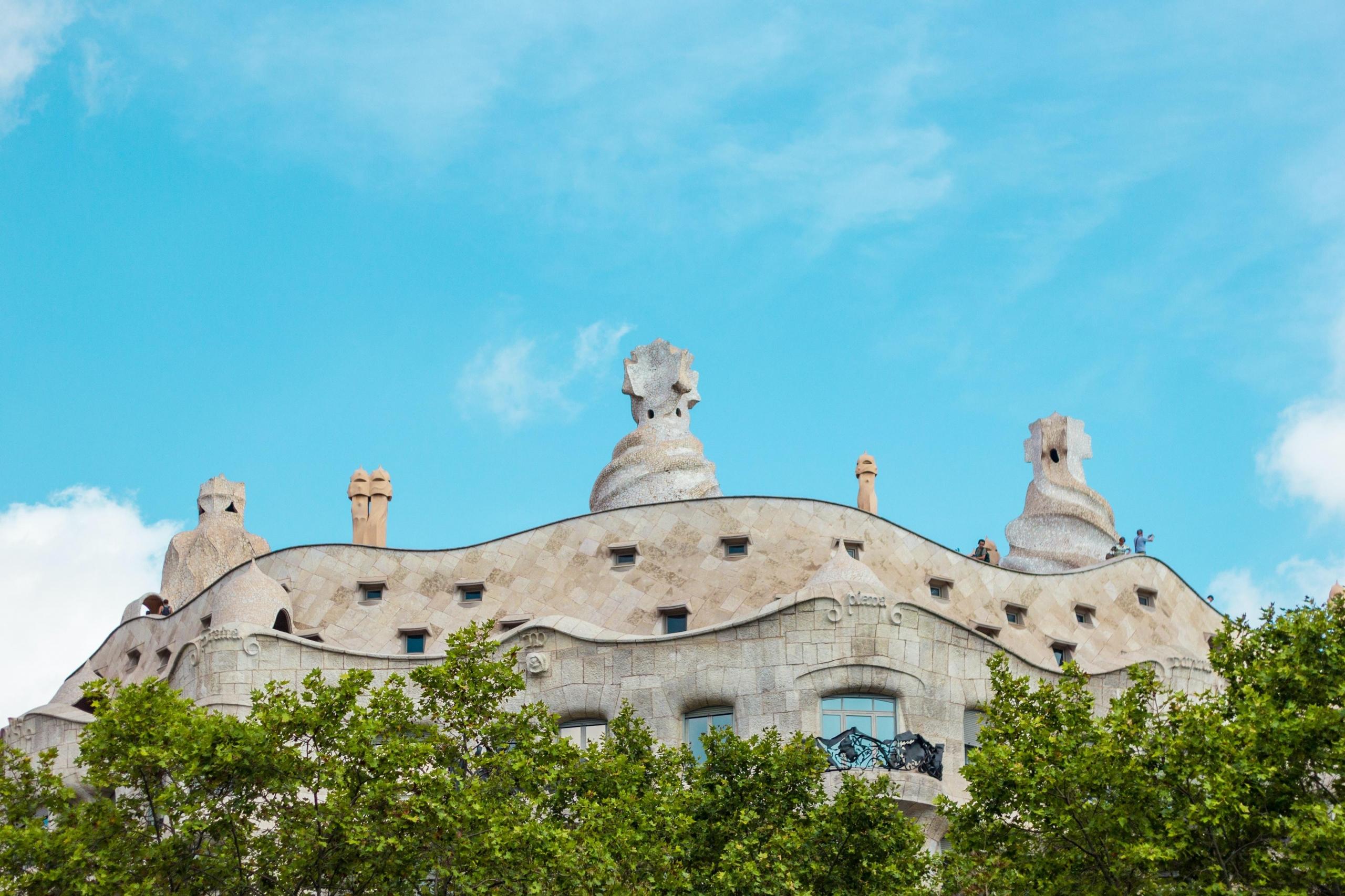 Bilde av bygning nedenfra, trekroner i forgrunnen, blå himmel i bakgrunnen (gaudi, casa mila)