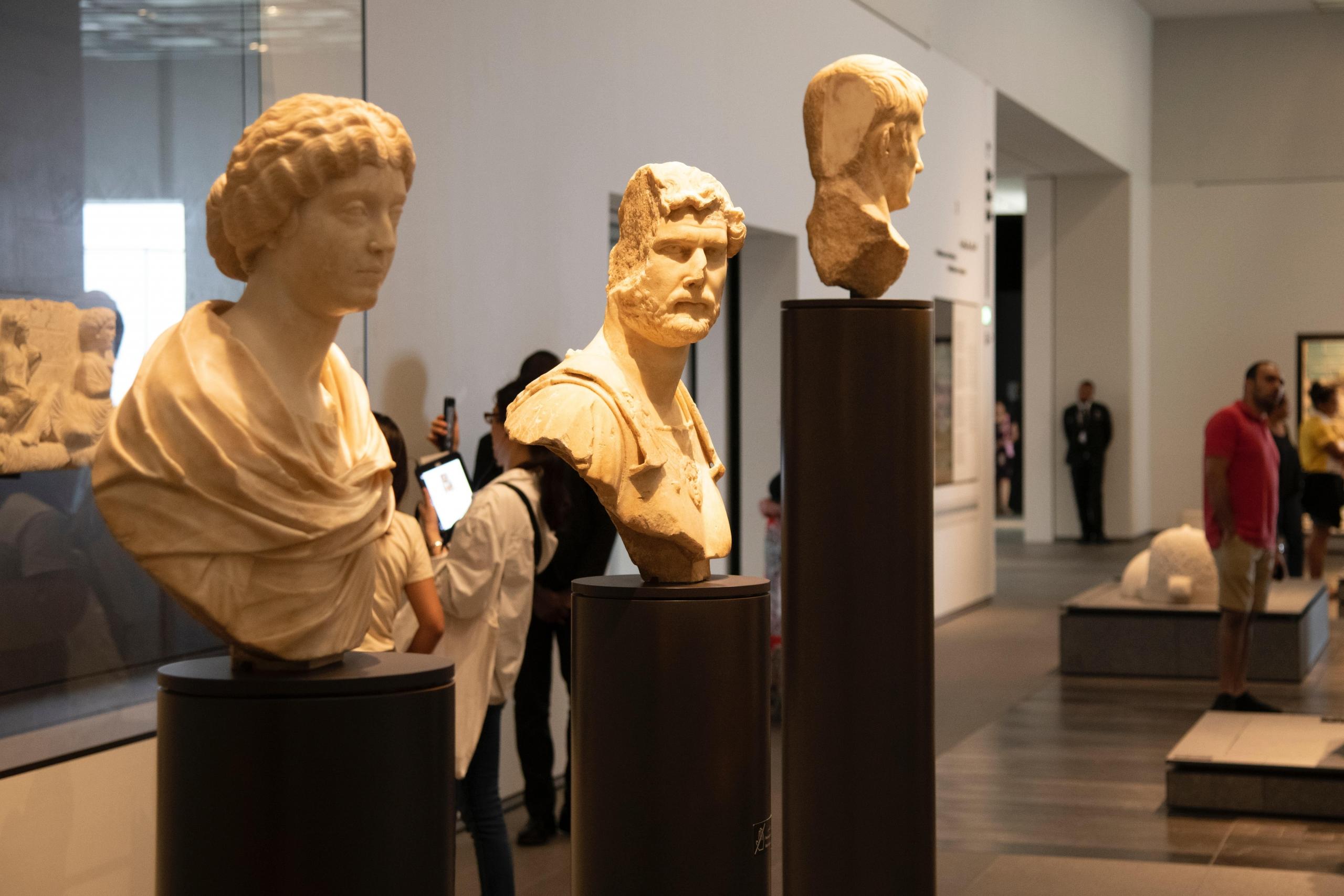 tre klassiske byster står i et museum, mennesker ser på kunst i bakgrunnen