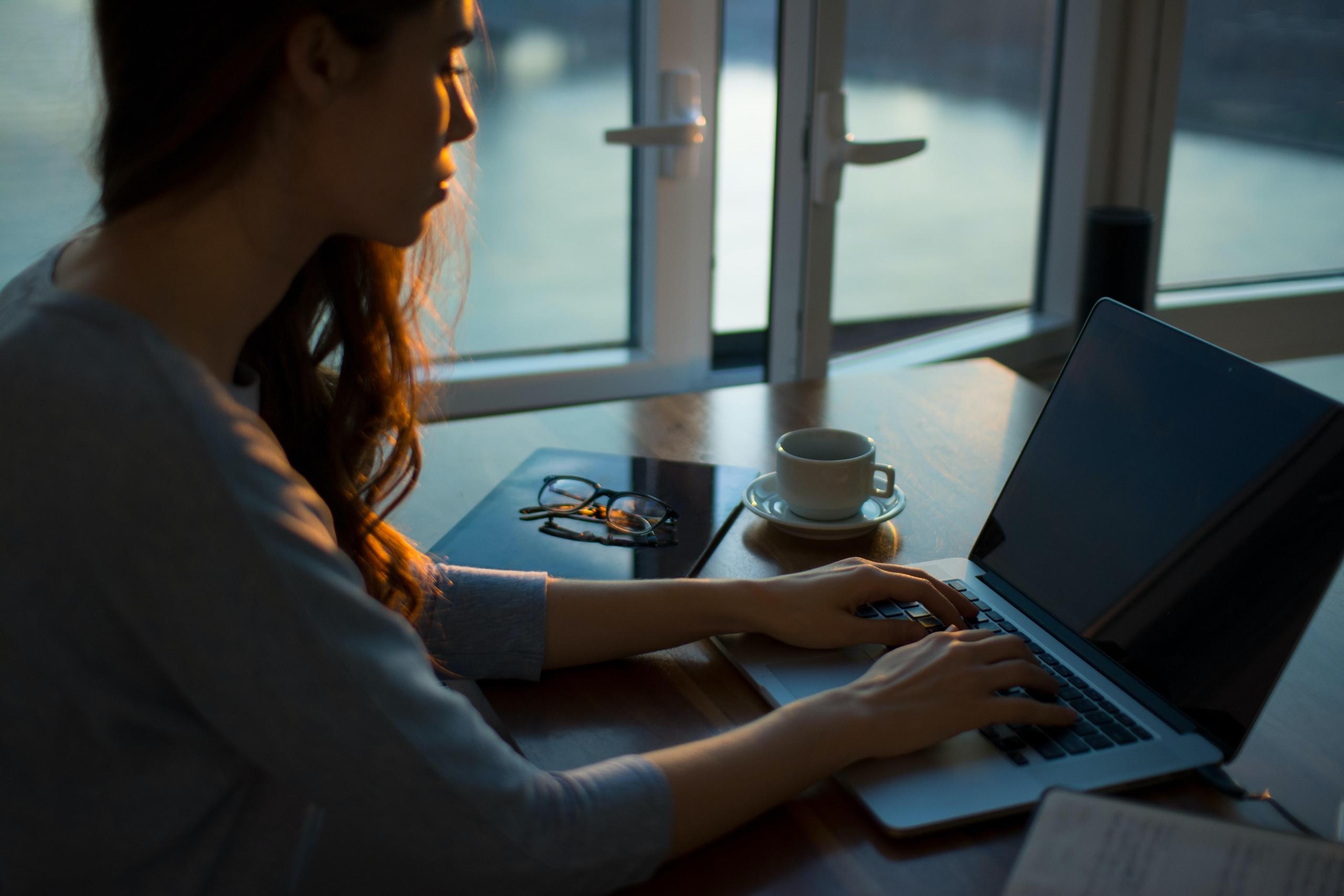 kvinne sitter i skumringsmørkt rom og skriver på en pc, det er notatbøker og en kaffekopp ved siden av henne
