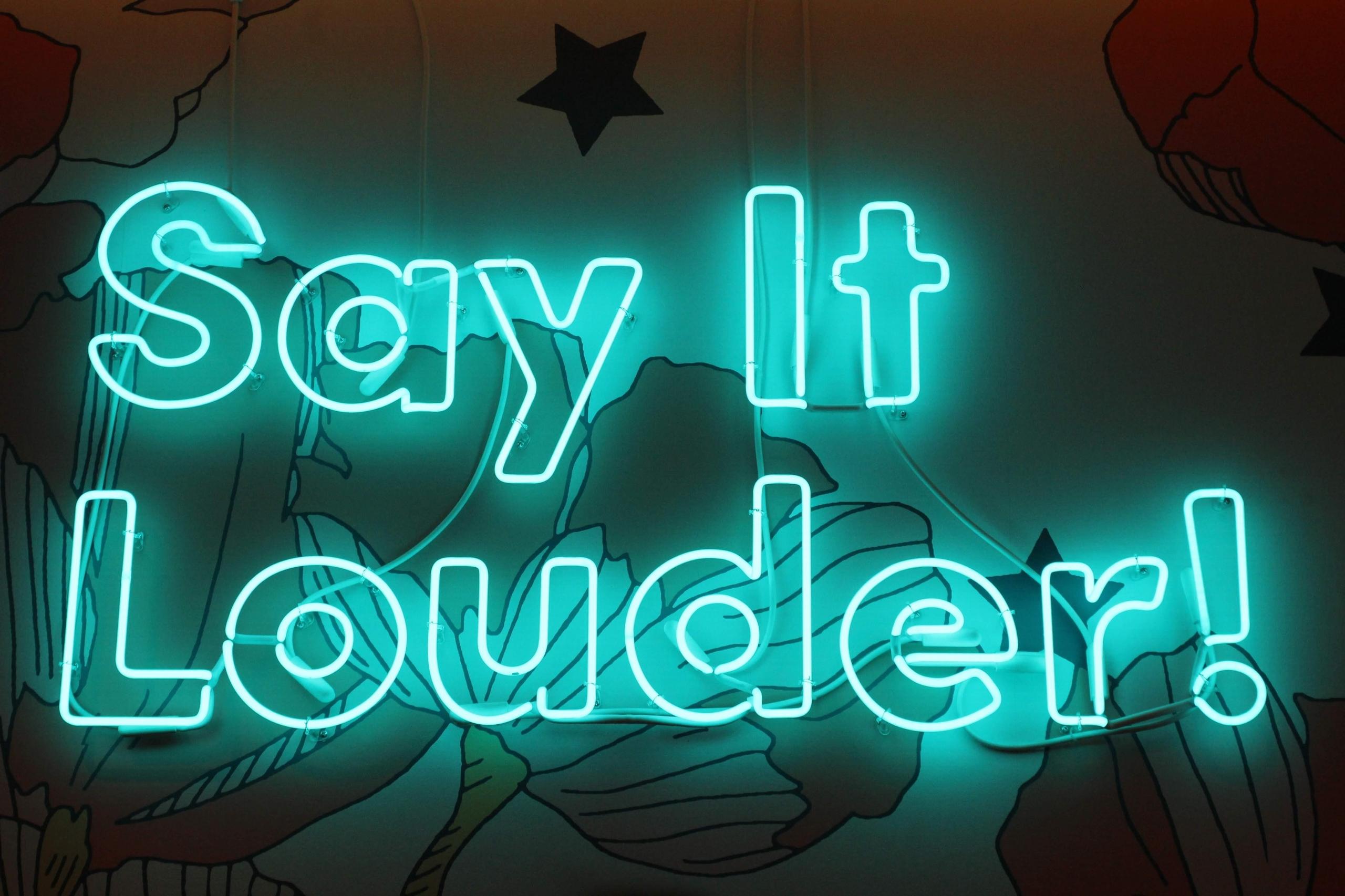 neonskilt på en vegg med blomstertapet, formet til setningen "Say it louder!"