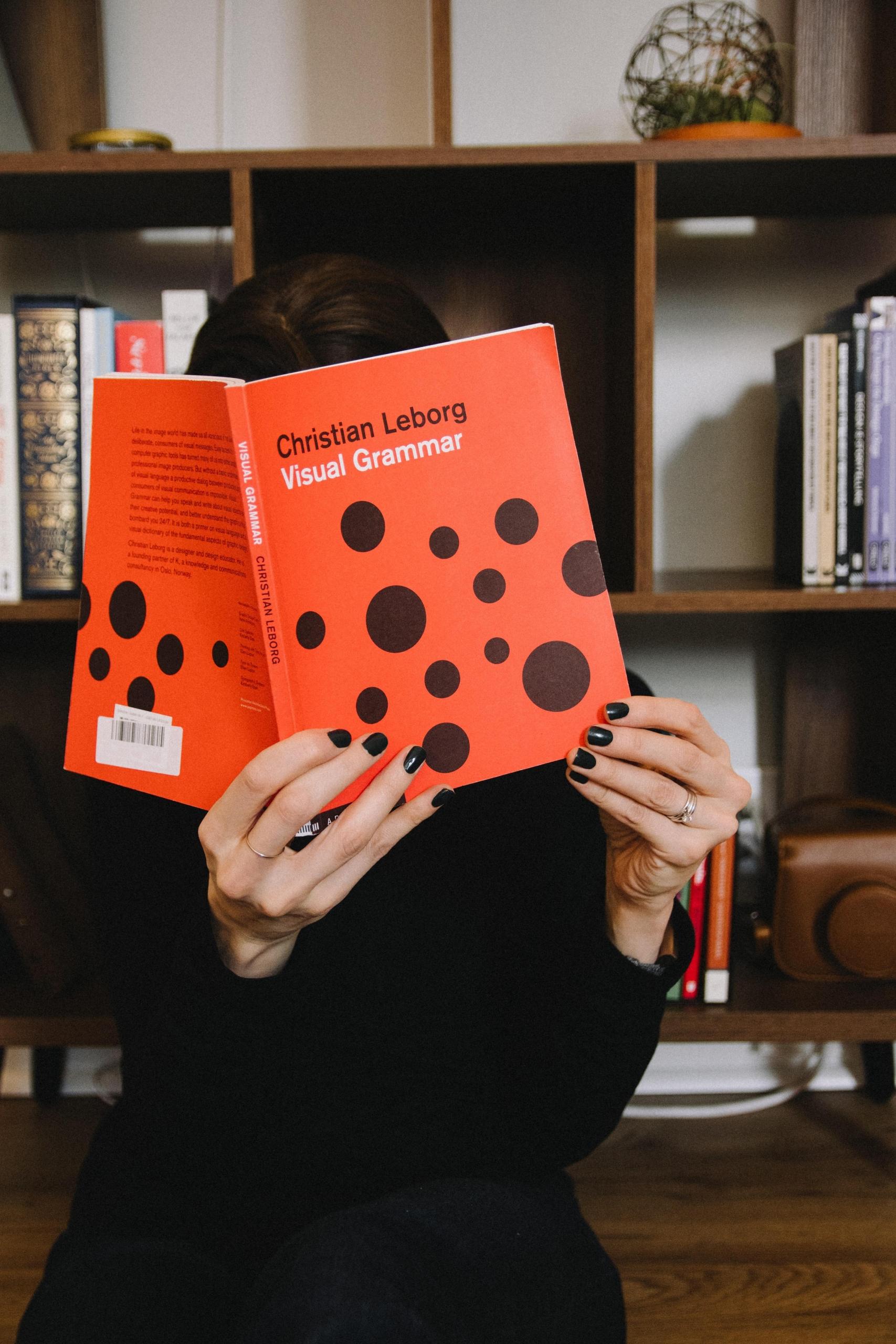 en person holder en bok som heter "Visual grammar" opp foran ansiktet, den er rød med svarte prikker, og man kan gjette at den har noe å gjøre med grafisk design eller lignende
