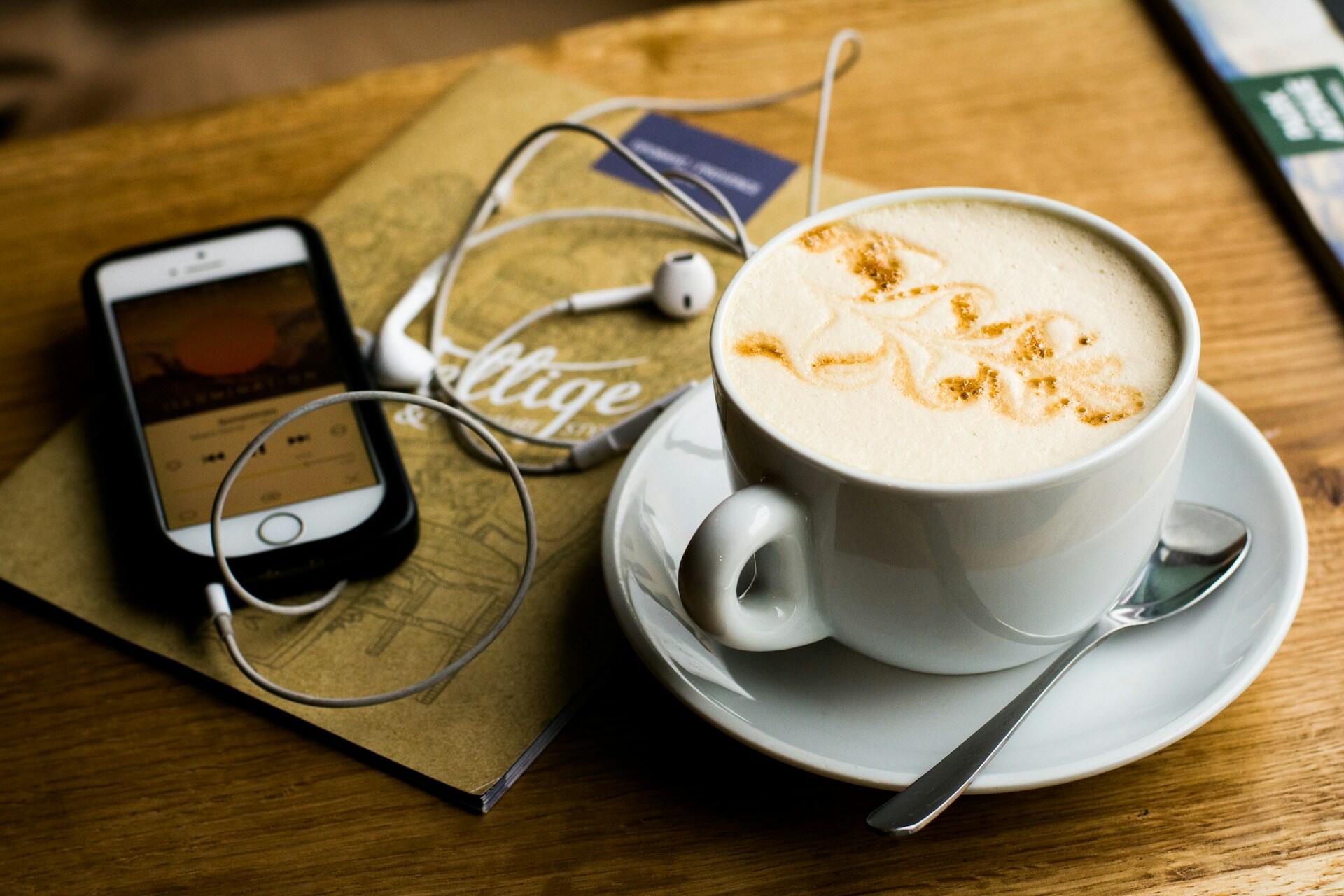 Kaffekopp og airpods tilkoblet mobil som lytter på podkast