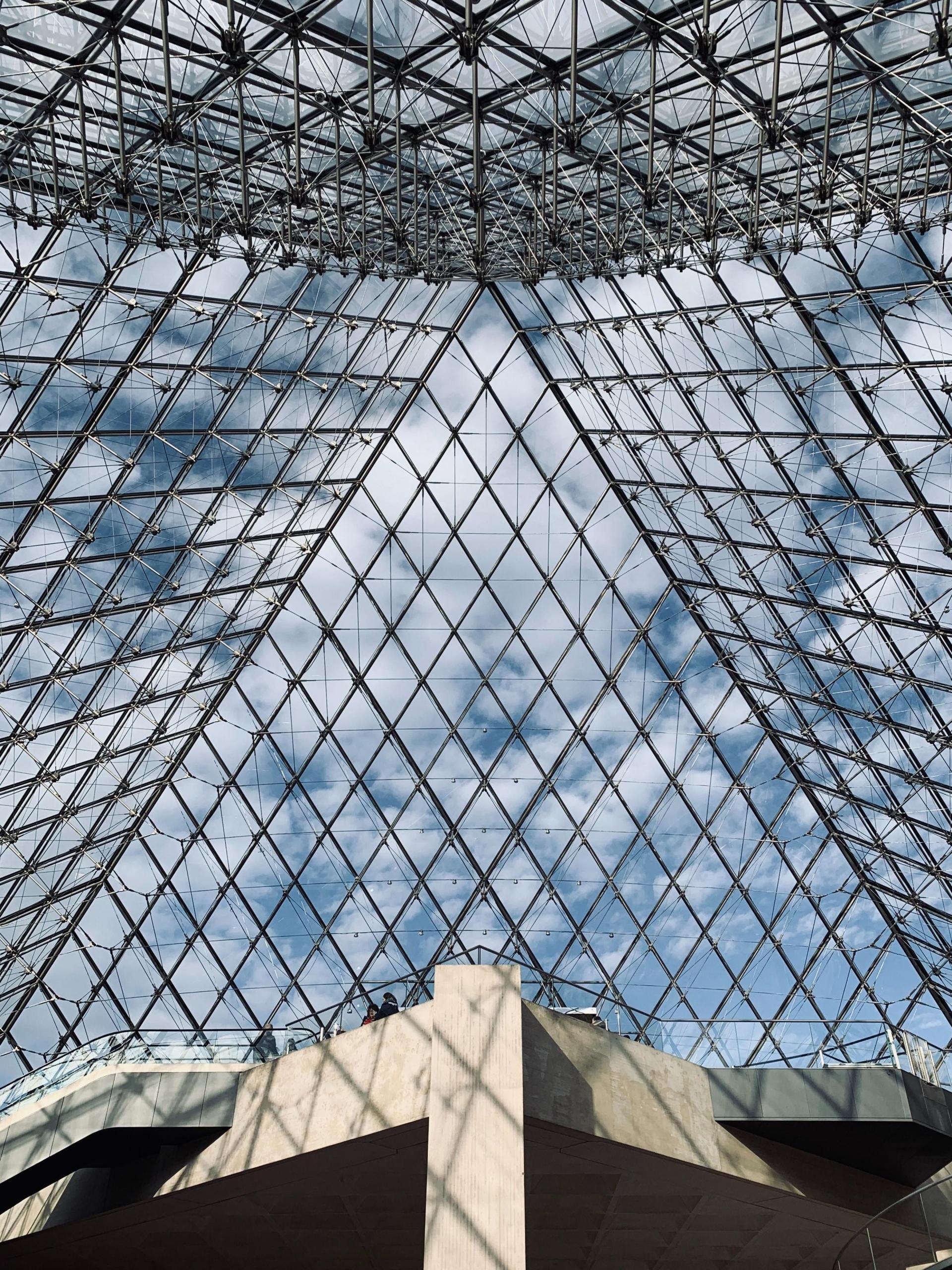 bilde nedenfra og opp mot innsiden av glasspyramiden ved louvre-museet i paris, gjennom glasset er himmelen blå med små, hvite skyer