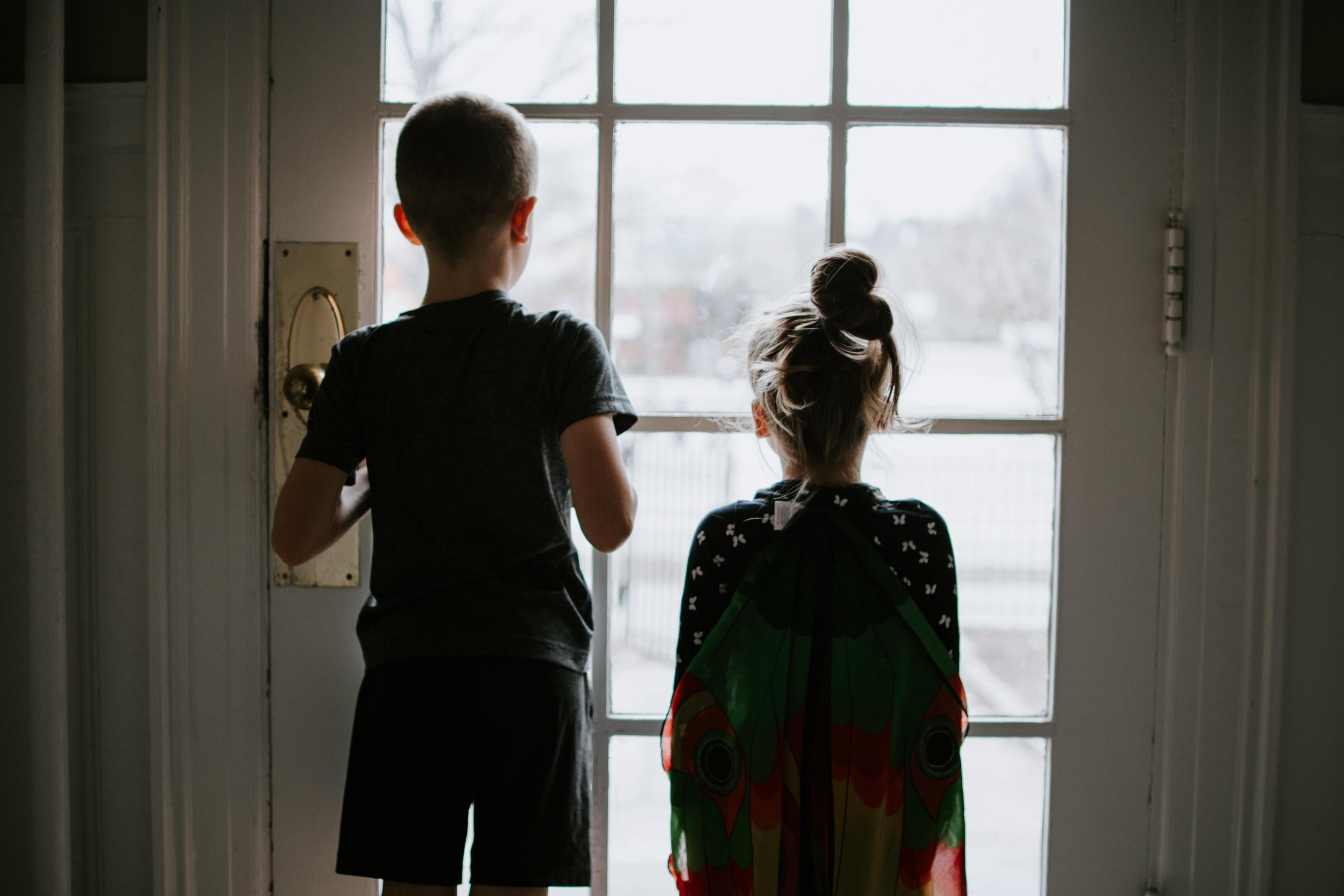 to barn, en gutt og en jente, står med ryggen til kamera i et halvmørkt rom og ser ut av et vindu