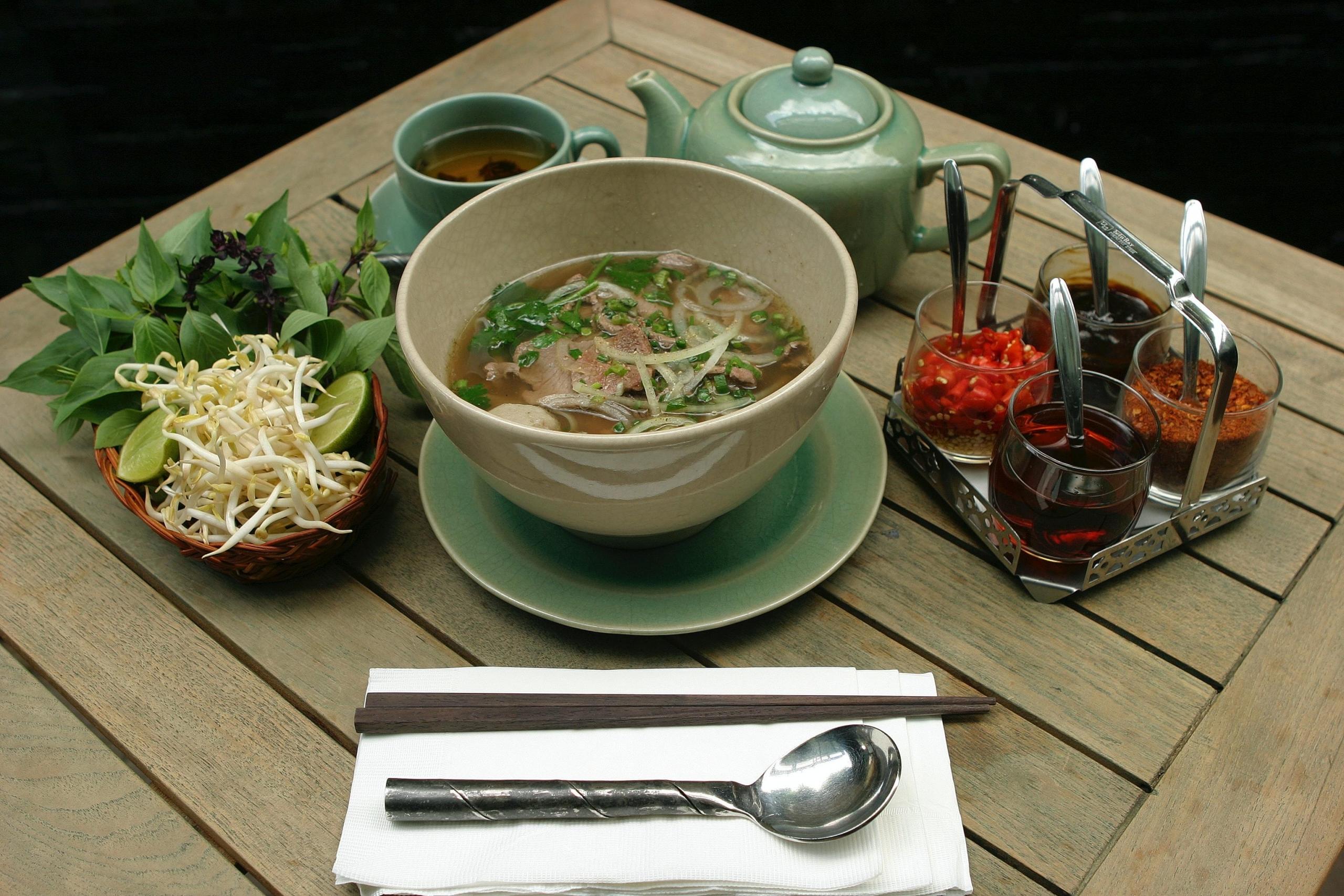 vietnamesisk pho med tilbehør på begge sider av bollen, på den ene noen sauser og hakket chili, på den andre spirer, urter og lime
