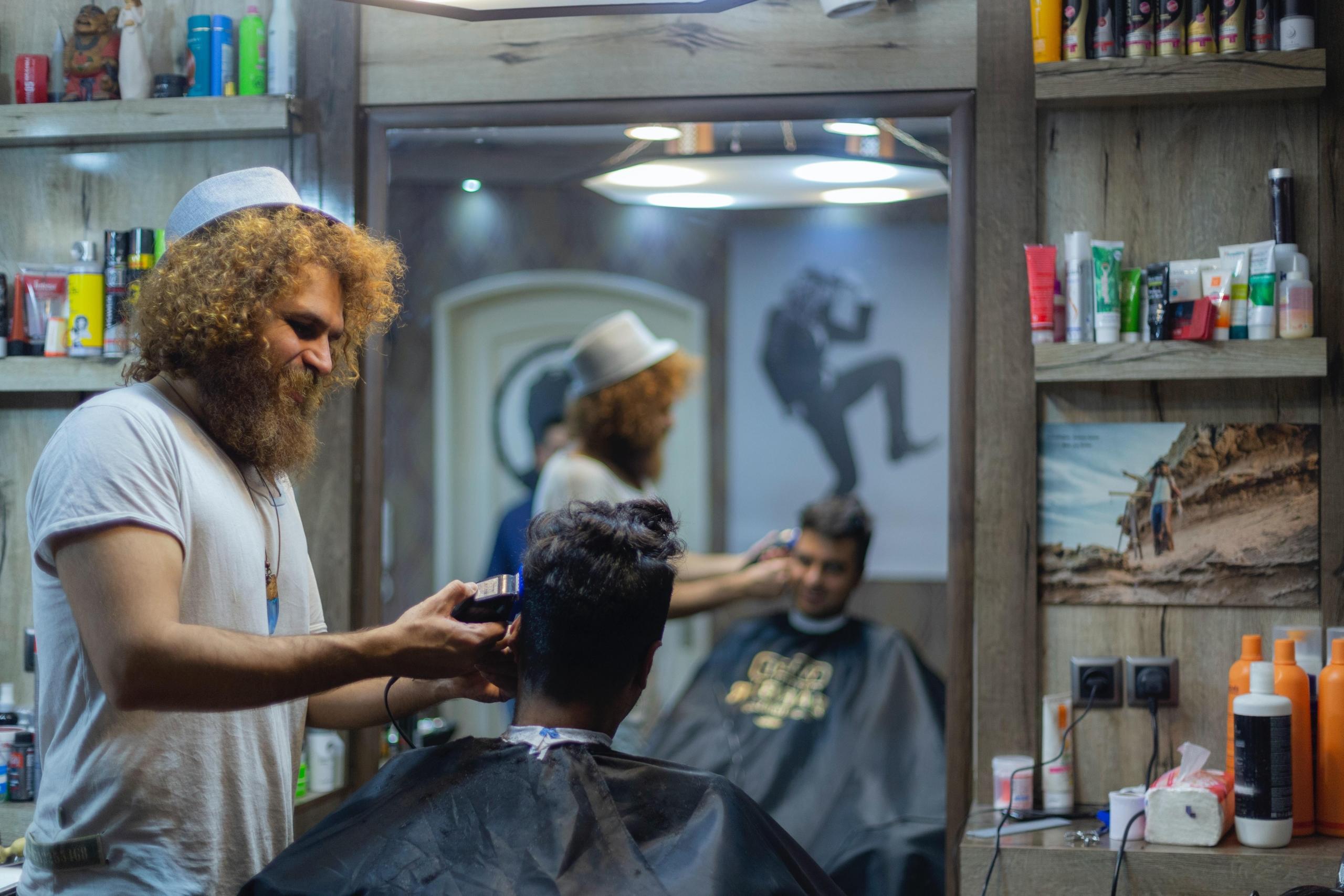 en mann med krøller i en salong holder en barbermaskin opp mot hodet til mannen som sitter i stolen, masse produkter står på hyller på veggen