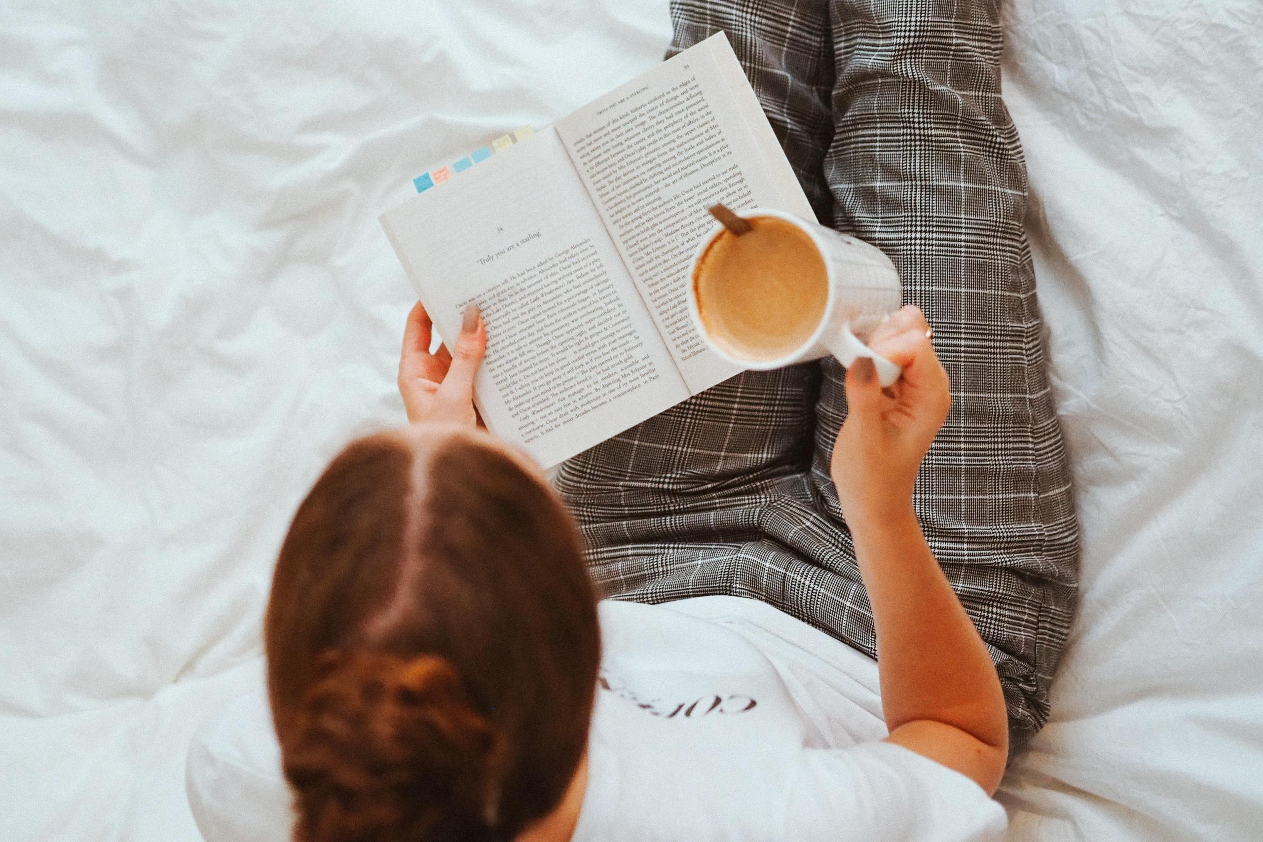 bilde ovenfra av en kvinne med rutete bukser som sitter og leser i det som ser ut som en seng. Hun holder en kaffekopp i den ene hånda
