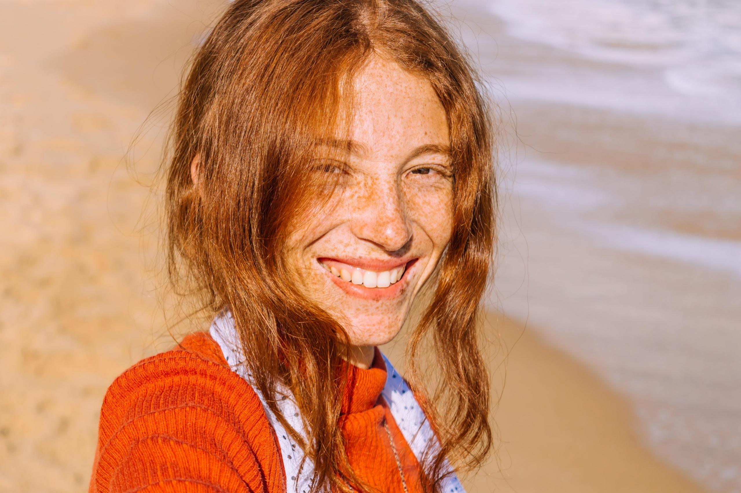 en kvinne med rødt hår og fregner smiler oppriktig til kamera, hun er på en strand