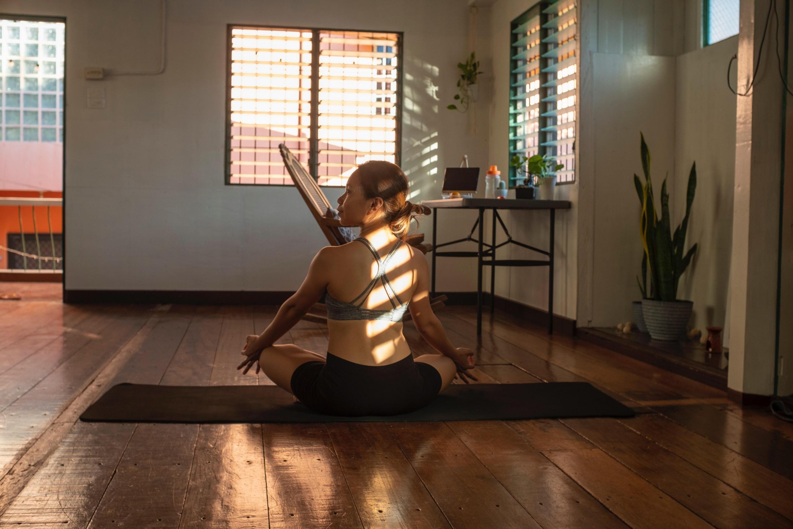 en kvinne sitter på en yogamatte i lotusstilling, sollyset treffer henne gjennom persiennene