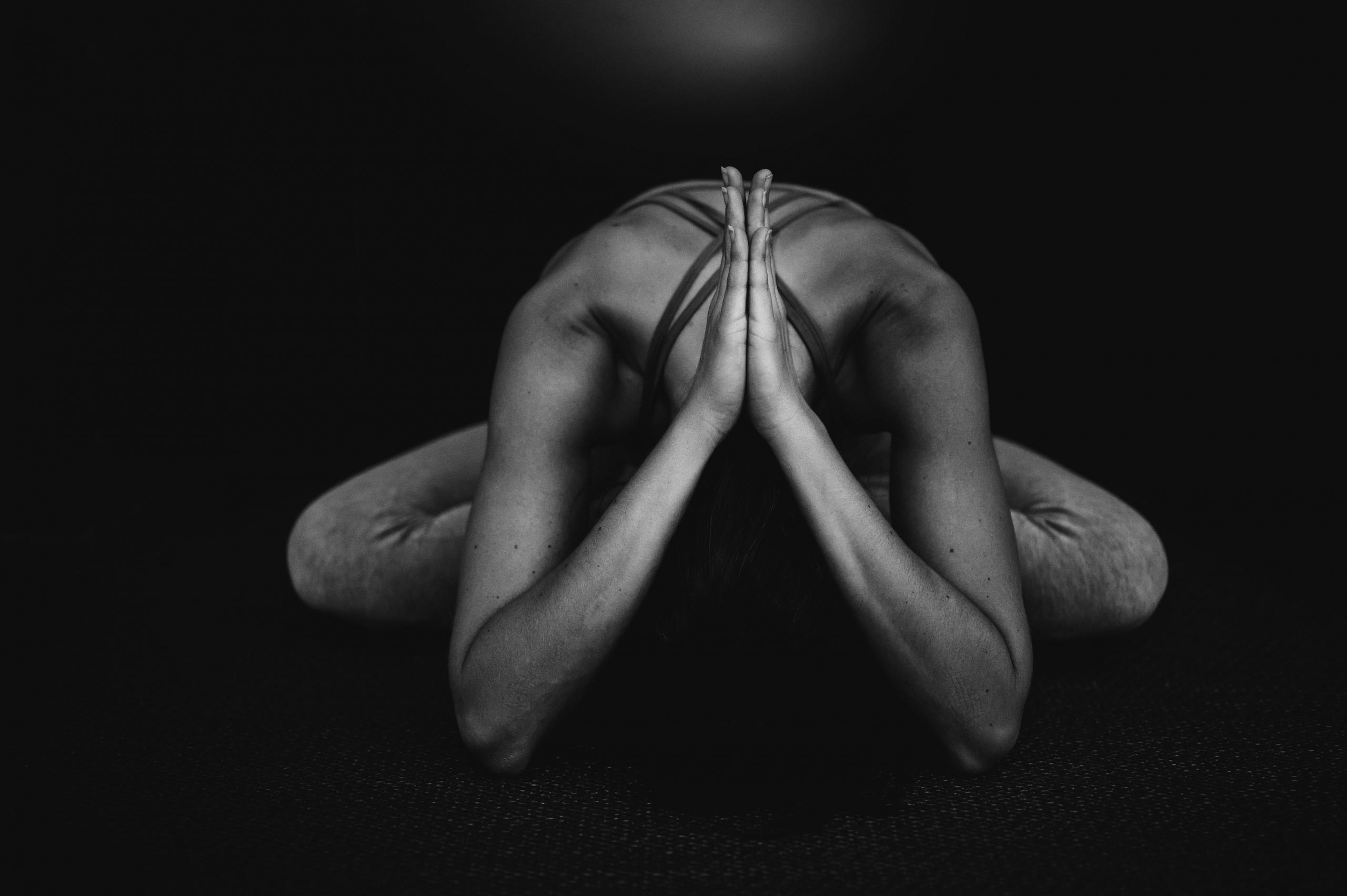 svart-hvitt-foto av en person i en sittende yogastilling, med knærne ut og føttene inn, albuene i gulvet og håndflatene samlet over hodet, som er bøyd mot bakken. Stillingen gir assosiasjoner til meditasjon