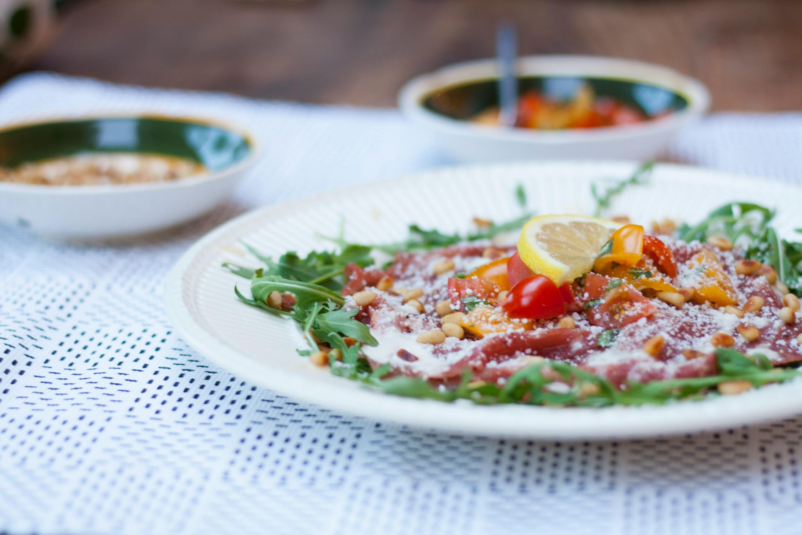 Et bord med hvit duk med tallerken med Carpaccio, et italiensk rett med grønnsaker og kjøtt. 
