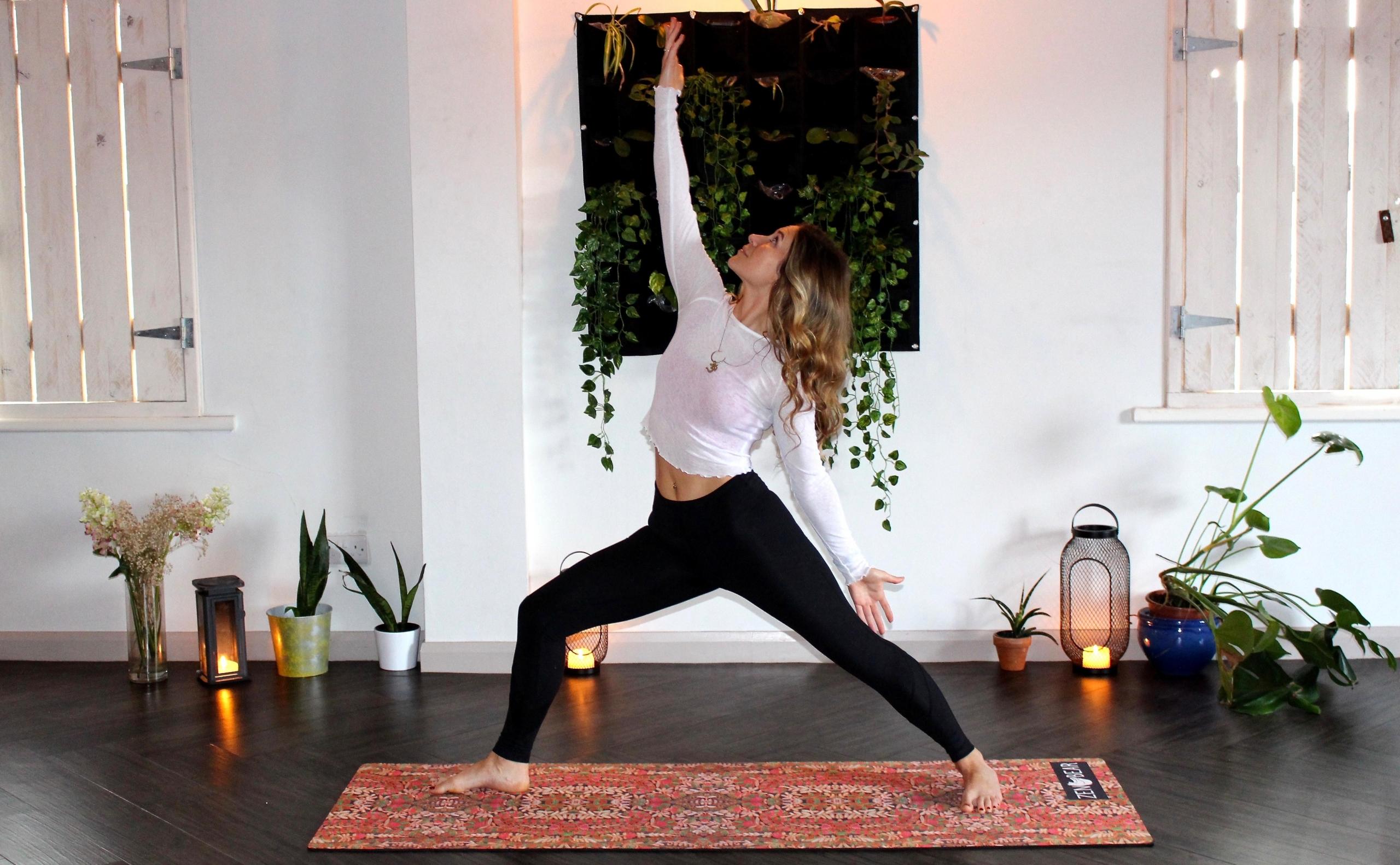 en kvinne driver med yoga i sitt eget hjem, hun står i en krigerstilling på en yogamatte og har på seg en svart tights og en hvit, tynn genser