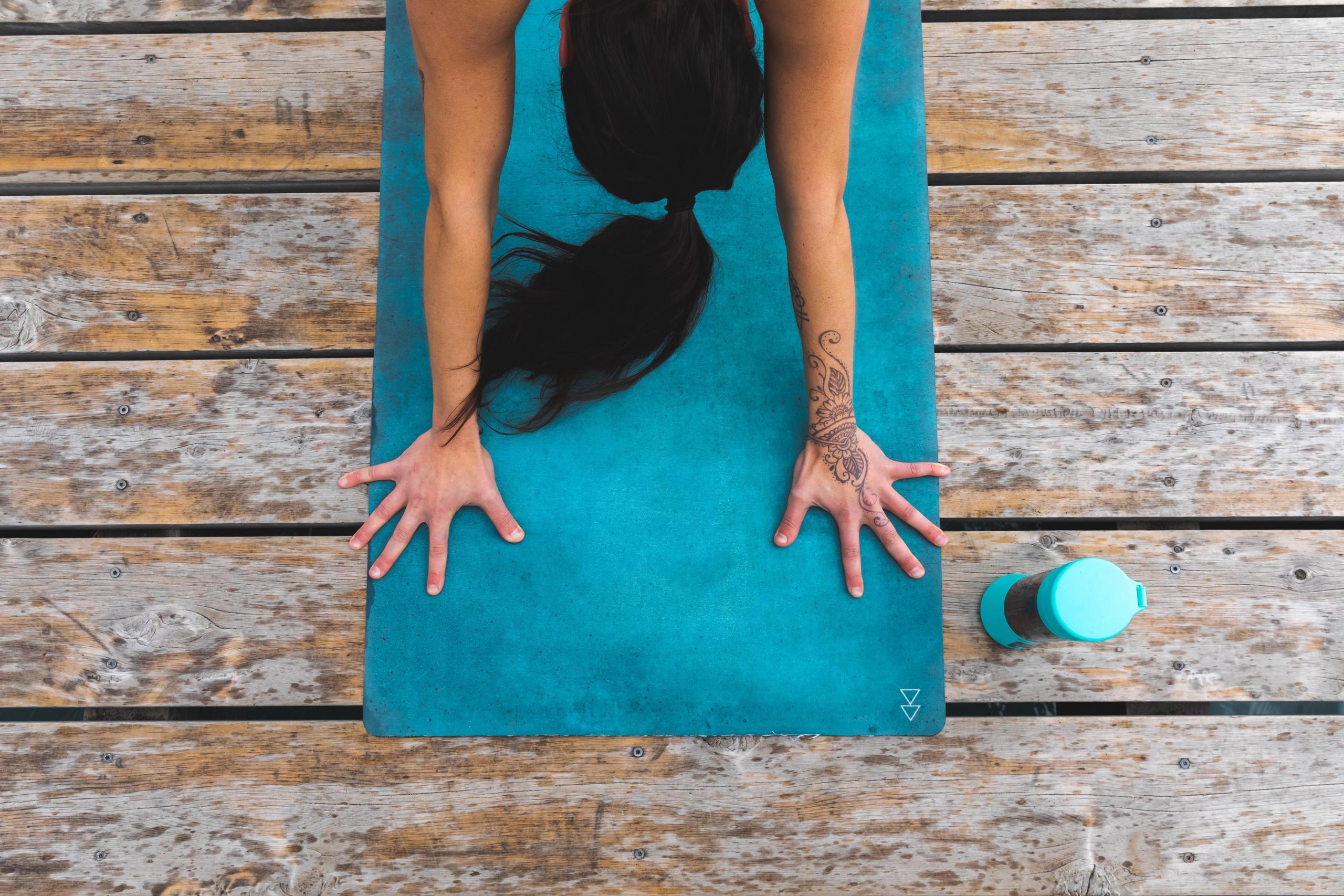 bilde ovenfra av en blå yogamatte og en kvinne som står i en yogastilling. Ved siden av står en drikkeflaske