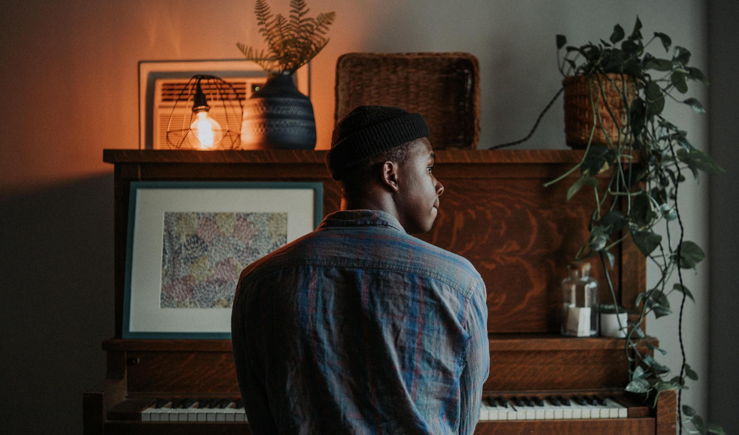 en ung mann sitter ved et piano i det som ser ut som hjemmet hans, det er mange dekorasjoner på pianoet, bilder og en lampe og en plante