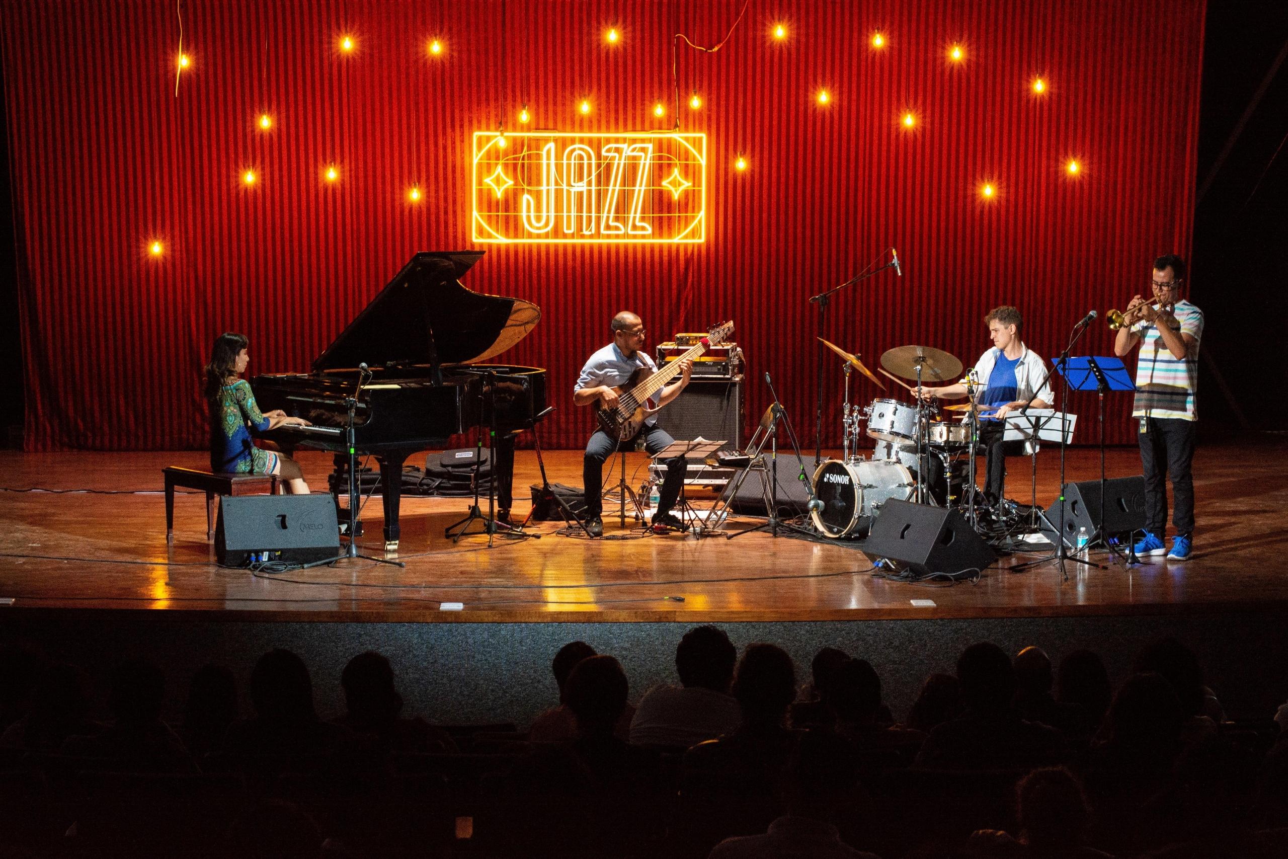 en gruppe på fire sitter på en scene med et neonskilt der det står "jazz", de spiller piano, trommer, trompet og et gitaraktig instrument med sju strenger