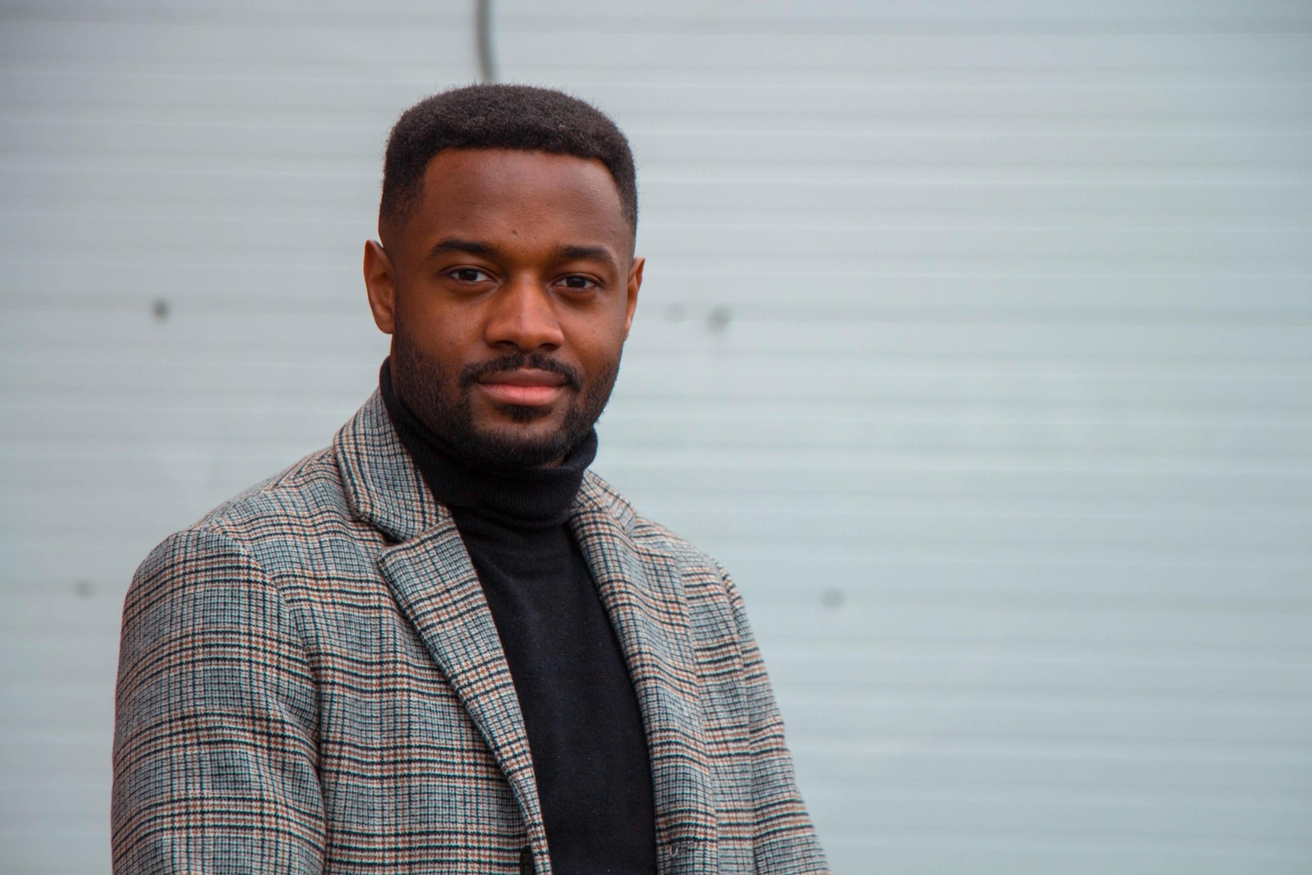 en ung mann i turtleneck og dressjakke ser i kamera og smiler avmålt