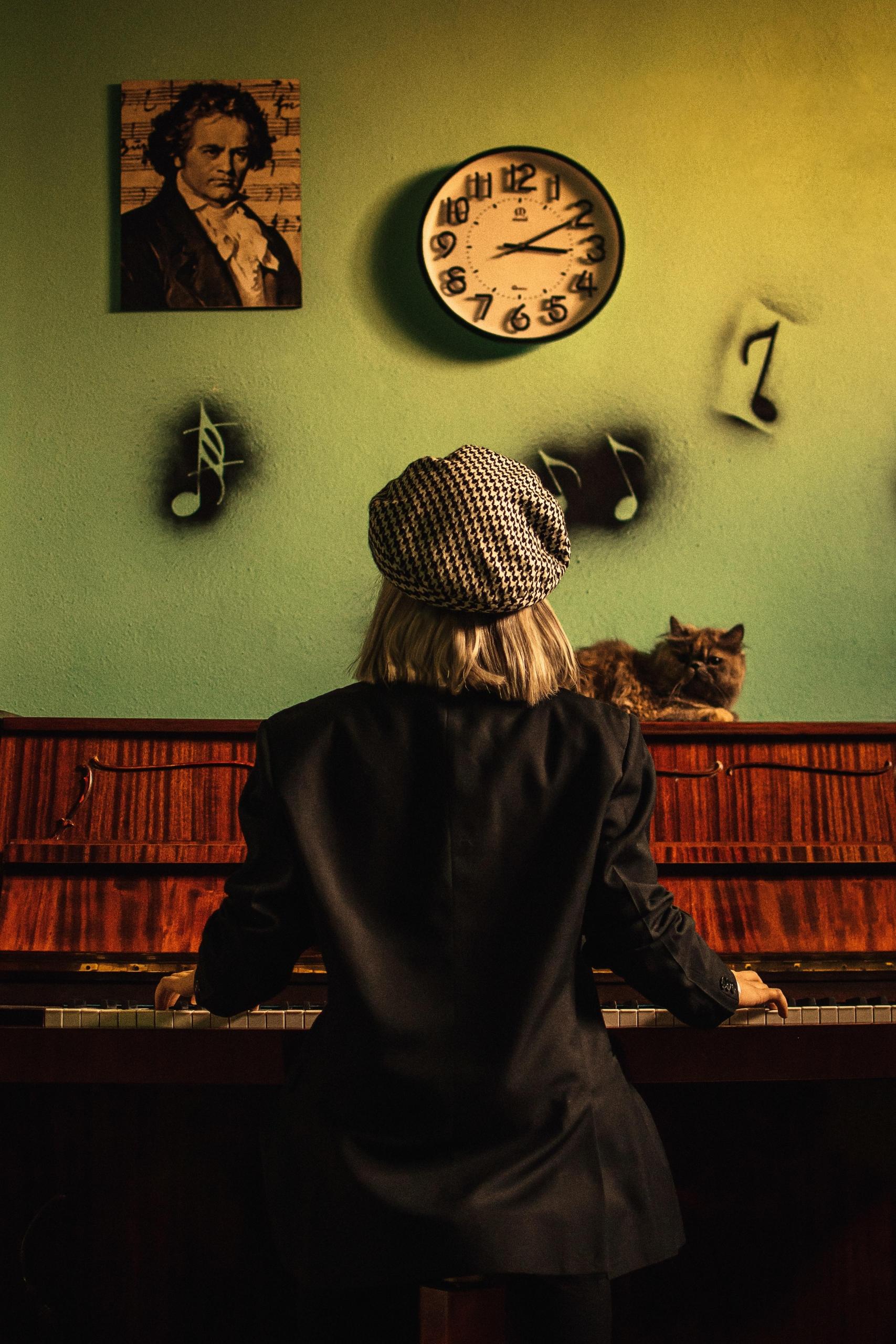 en person med dressjakke og sixpence-aktig hatt spiller på et piano med rød fløyel på innsiden, det henger et bilde av beethoven på veggen, og det er spraymalt noter på veggen