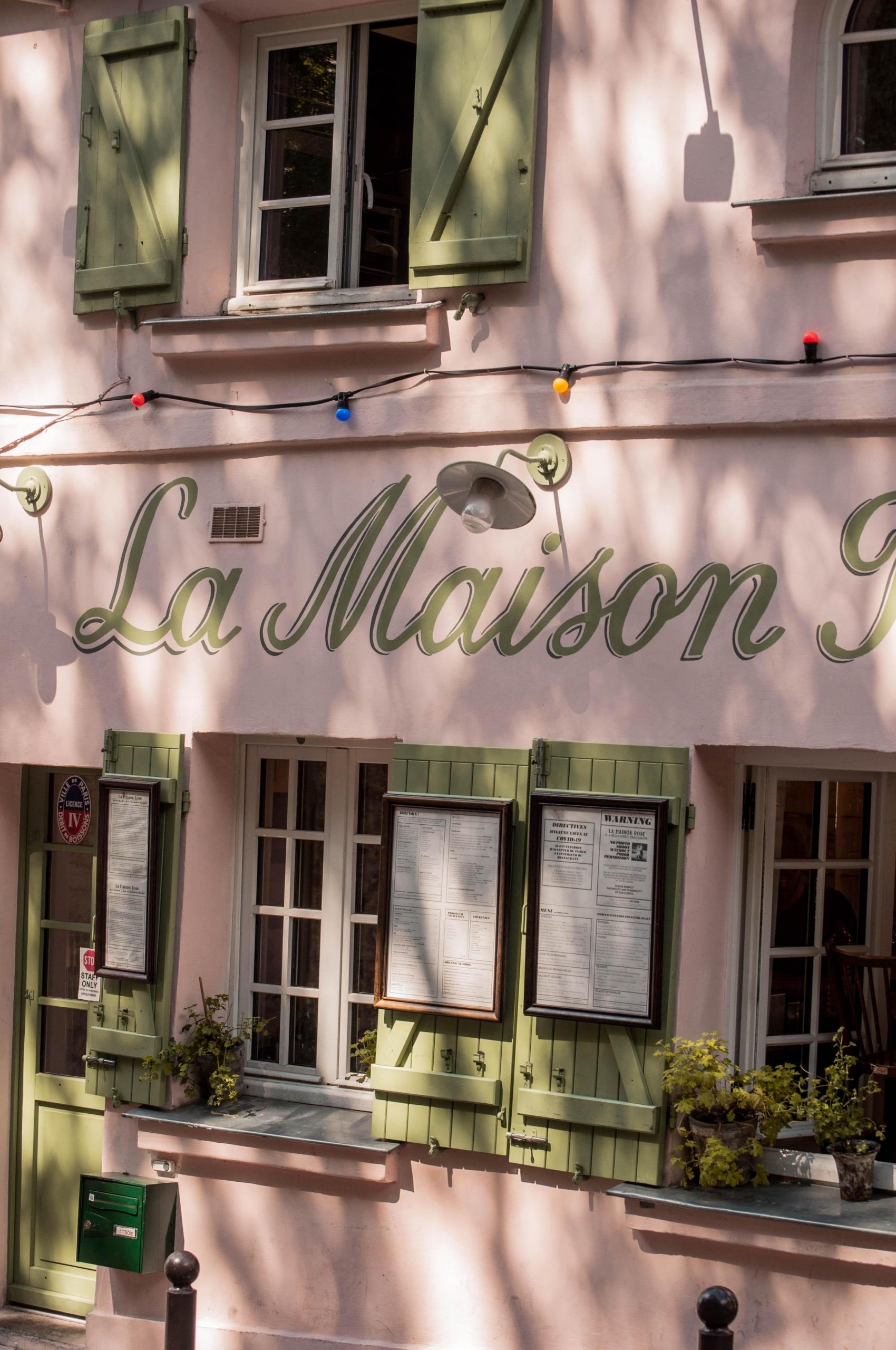 et rosa hus som det står "La Maison" på, vinduene har grønne skodder, og det henger menyer på dem