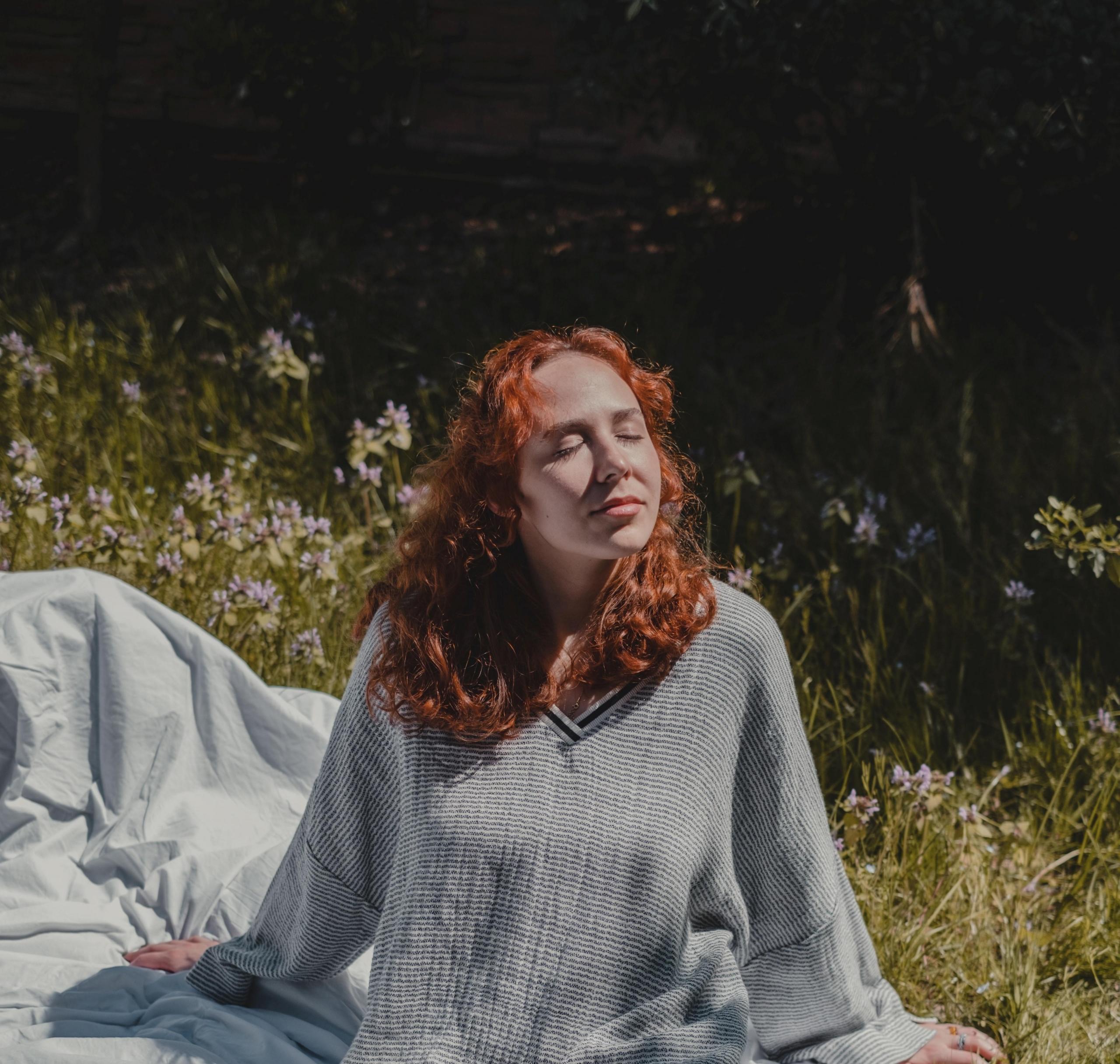 en jente sitter på et teppe på gresset i sola, hun har hodet vendt opp og øynene lukket, og smiler forsiktig