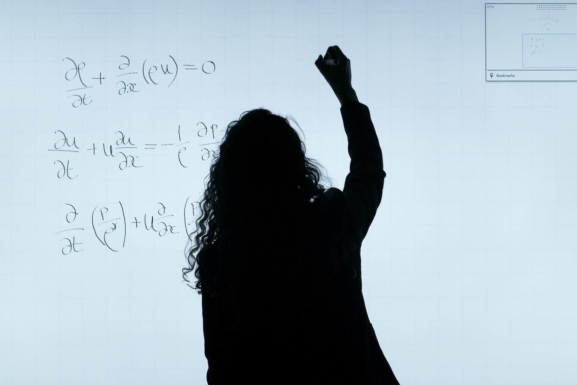 Kvinne står med ryggen til og skriver matematiske stykker på en lystavle.