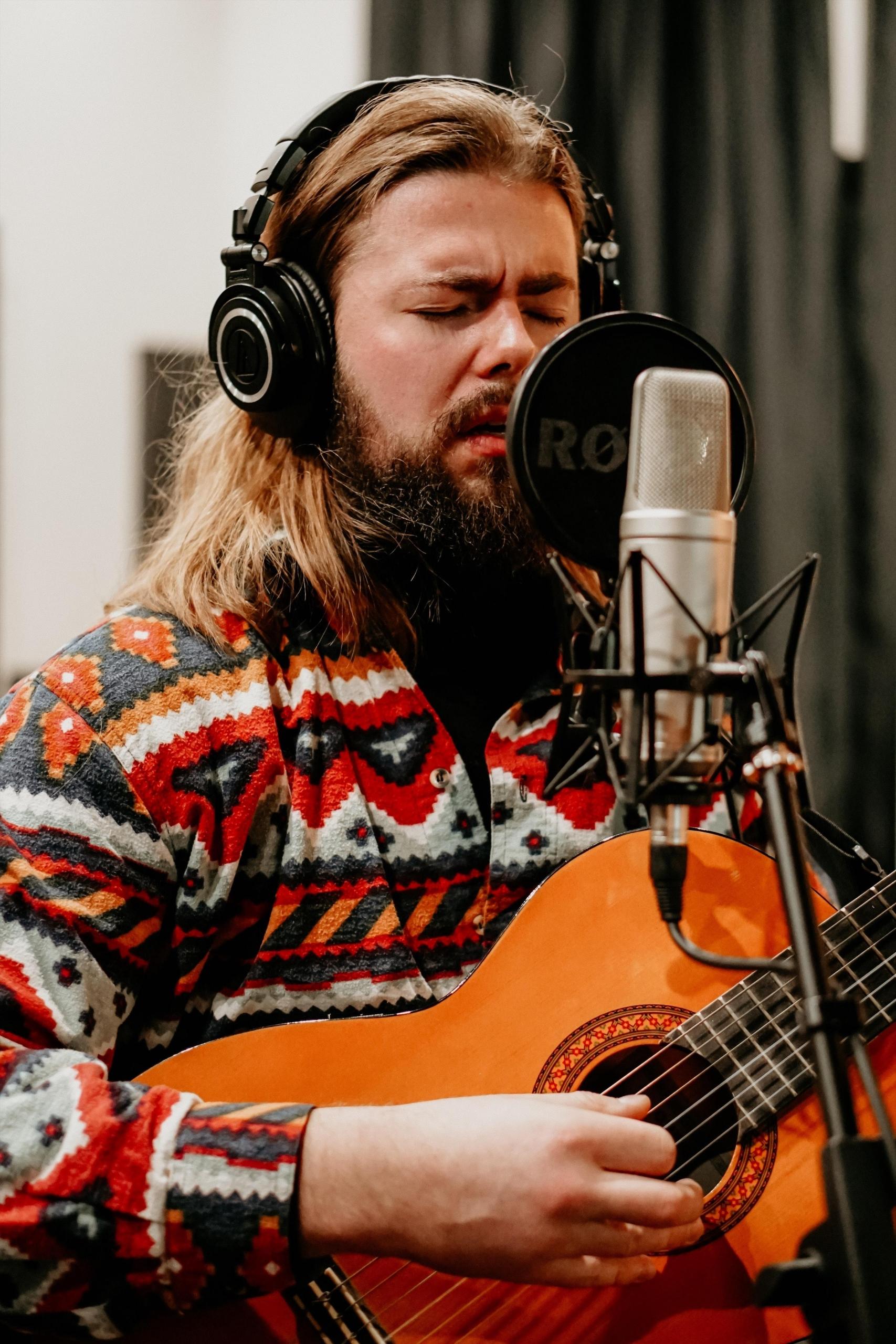 en ung mann synger inderlig inn i en mikrofon i et studio, kan spiller på en akustisk gitar