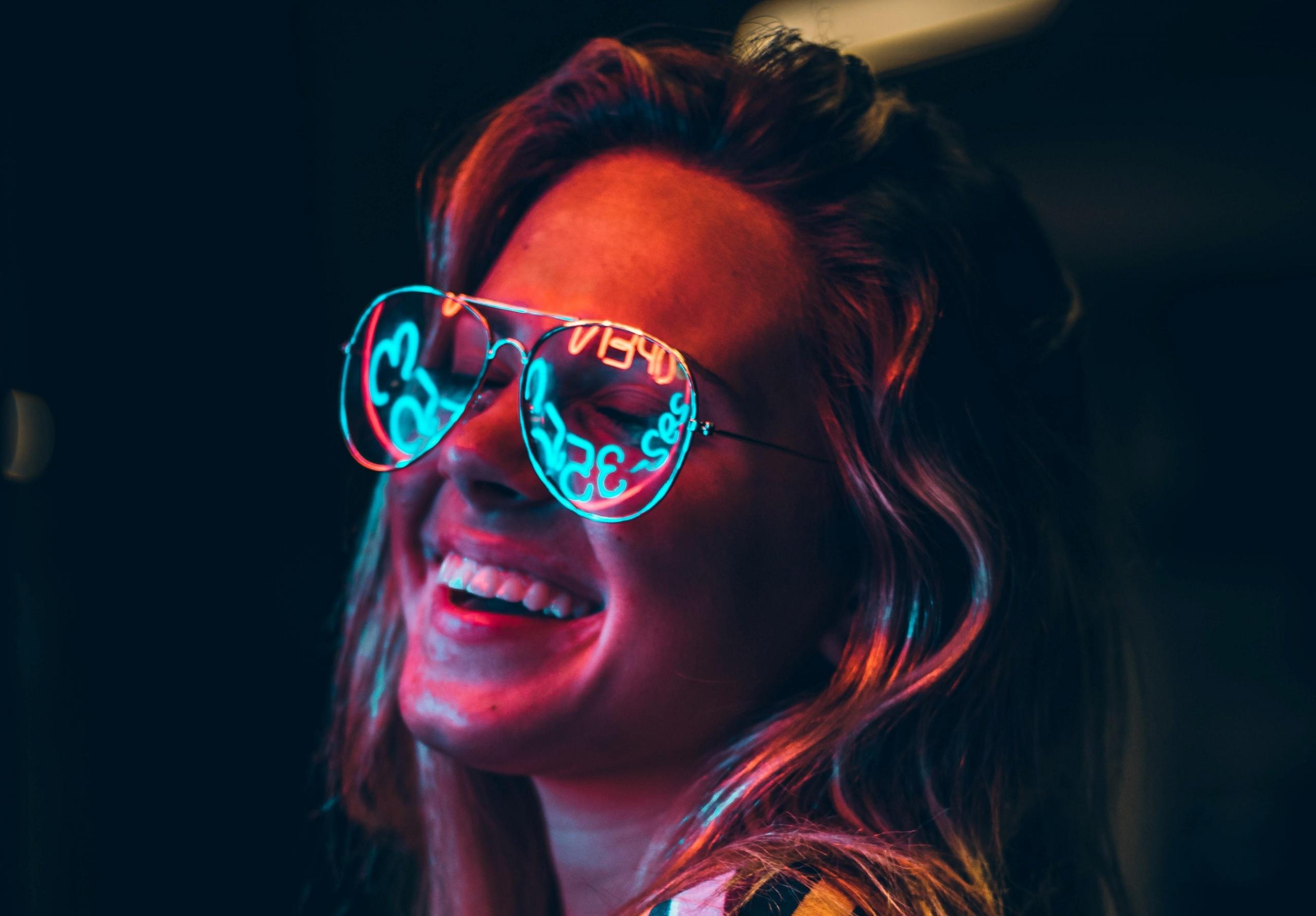 smilende kvinne i neonbelyst natt har på seg briller som reflekterer tall og bokstaver