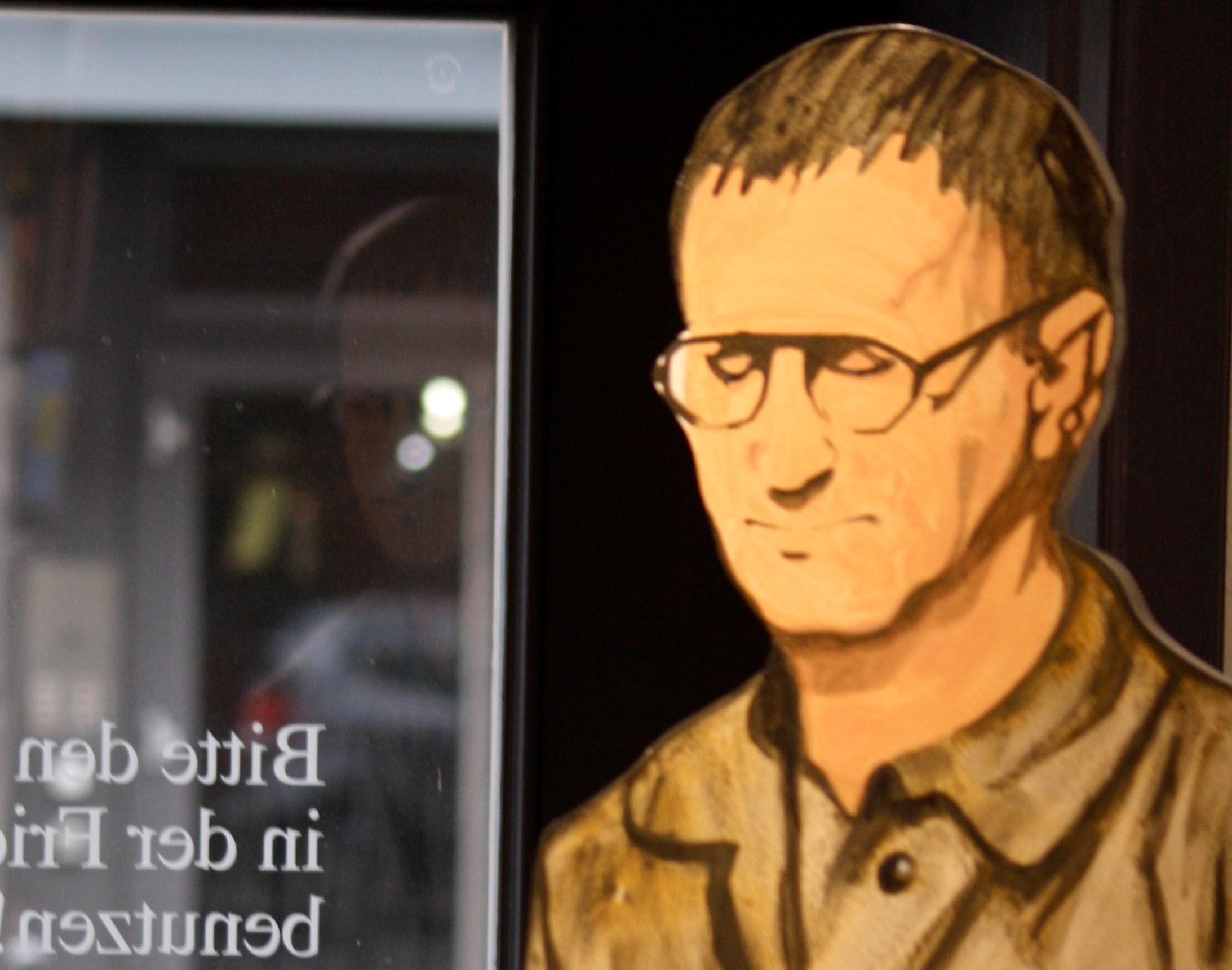 stor tegning av brecht på veggen i et offentlig rom, en kafé eller lignende, l bakgrunnen er et vindu ut mot gaten og gjenskinnet av en mann som ligner merkelig på tegningen