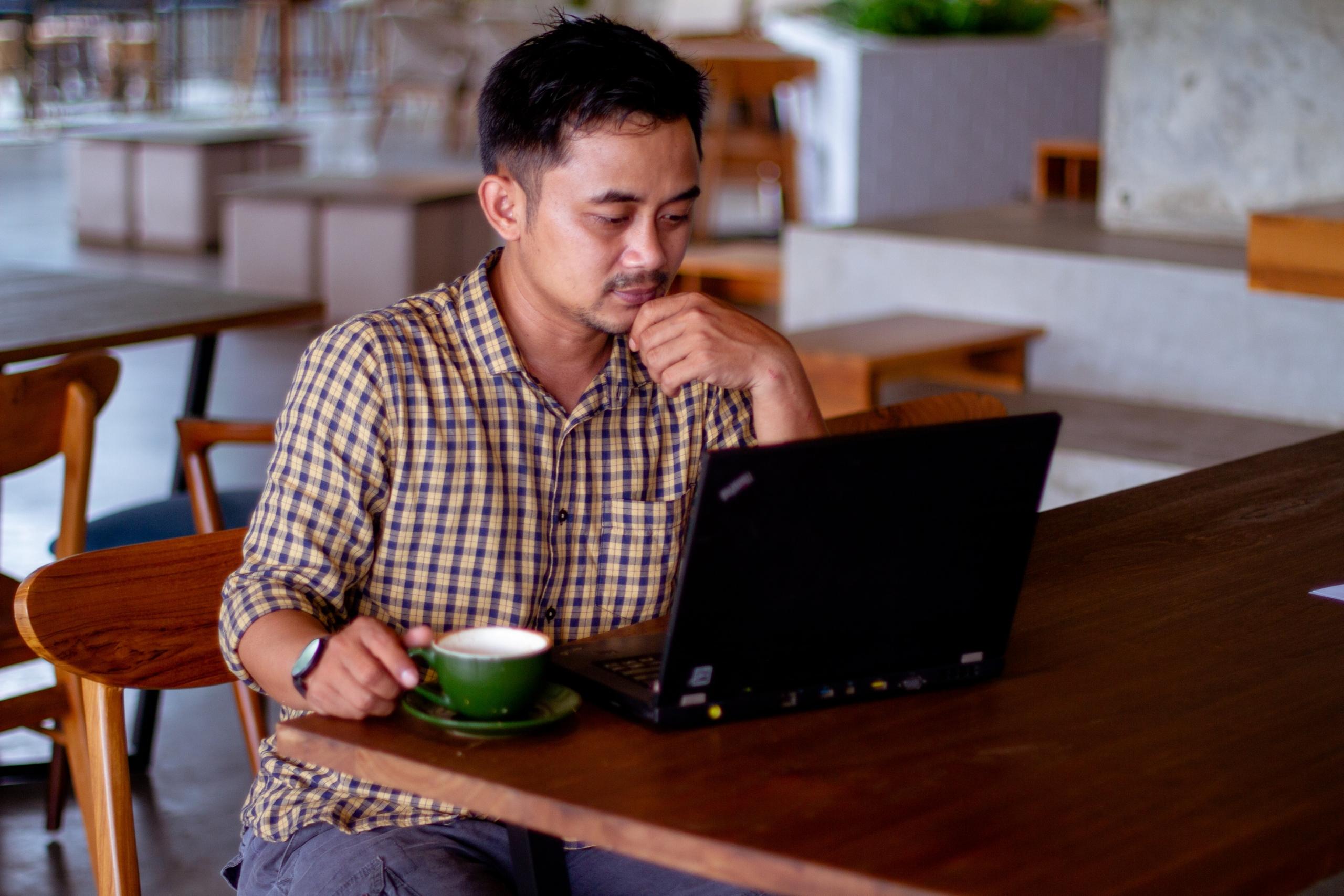 en mann sitter med en laptop og en kopp kaffe i en tom kafé, han ser tankefullt på skjermen