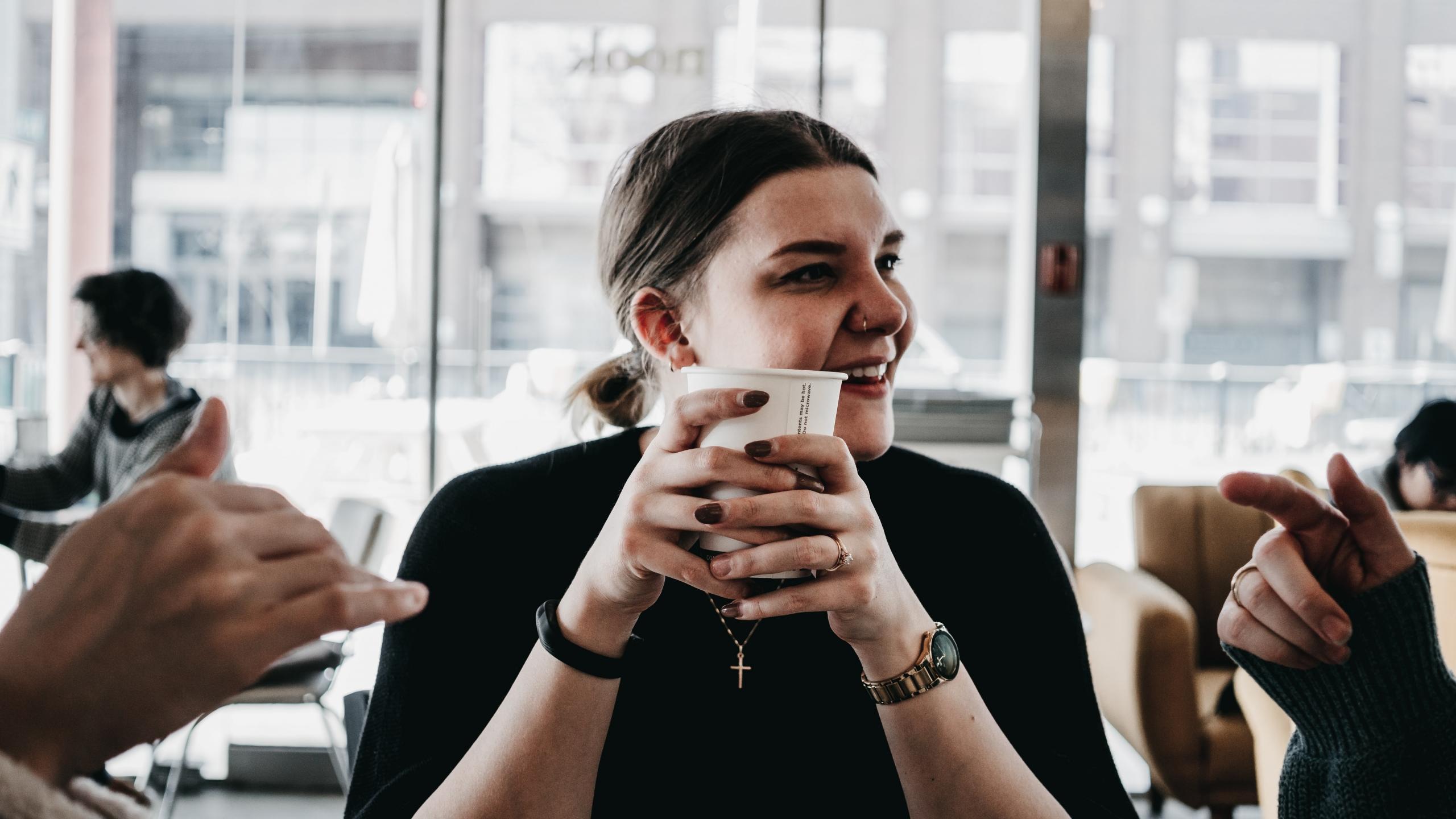 en jente sitter ved et bord og holder en pappkopp, hun smiler og ser til siden, det er tydelig at hun sitter sammen med noen fordi hendene deres stikker inn i bildet