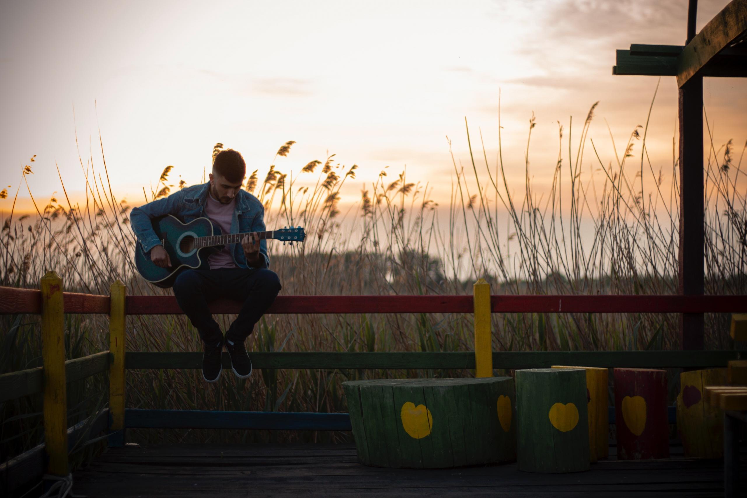 en mann med gitar sitter ute på et gjerde i solnedgangen