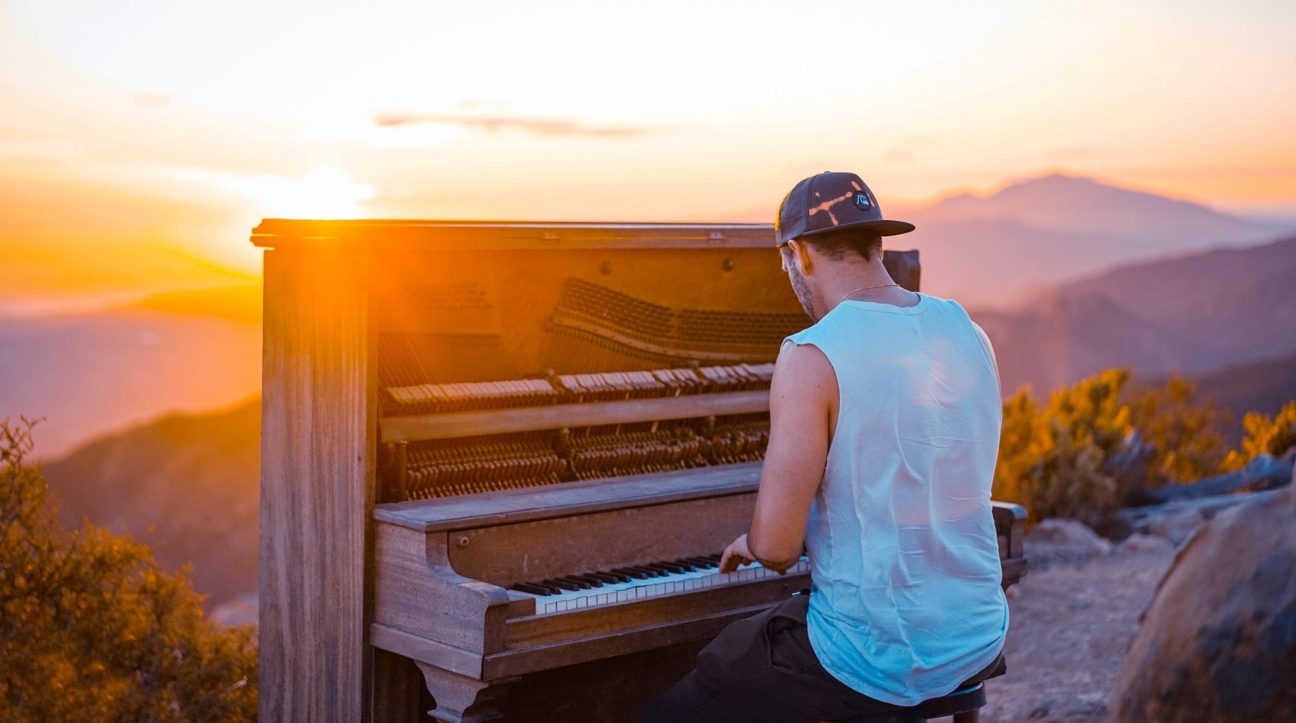 en mann spiller piano på det som ser ut som en fjelltopp, sola går ned i bakgrunnen