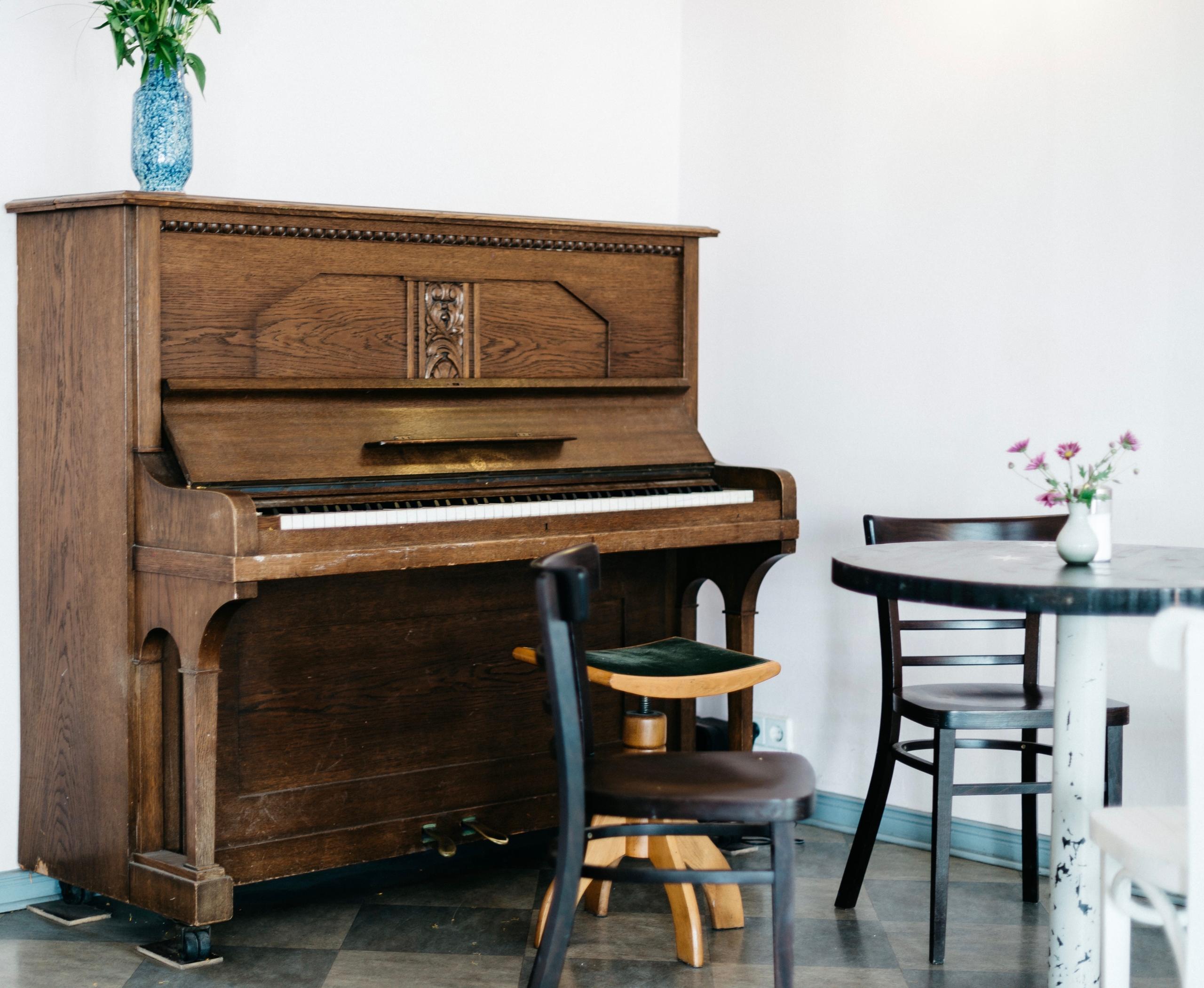 Et piano står inntil en vegg med en pianokrakk foran, den kan justeres ved å spinnes, det står et bord med stoler i annen høyde rett bak