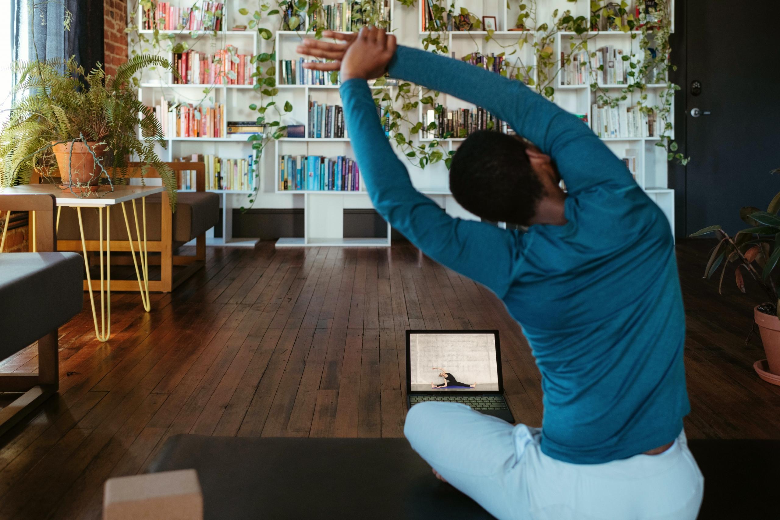 en mann sitter i stua si og følger en yogaøkt på laptopen sin