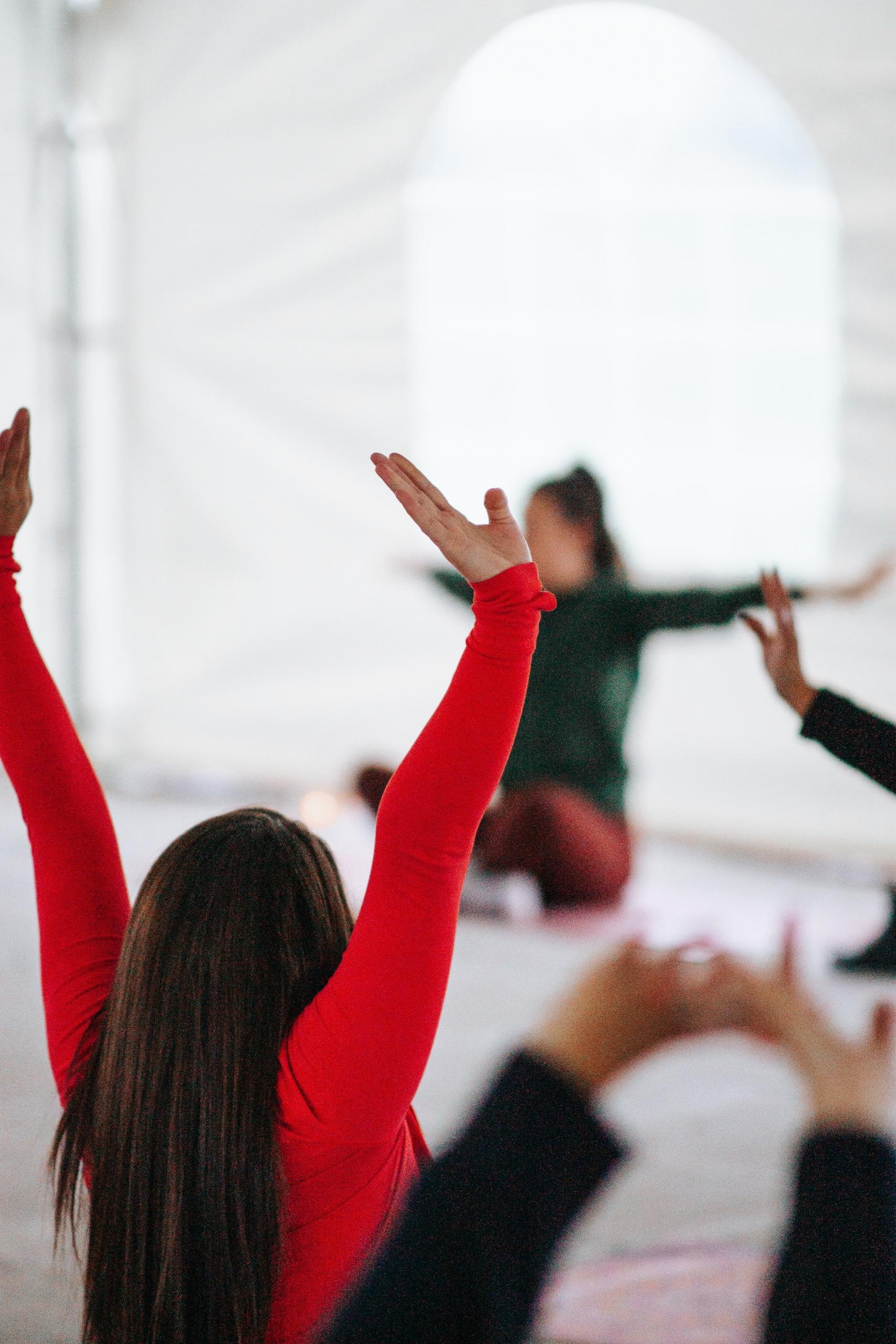 kvinner sitter i et stort hvitt studio på yogamatter, de har beina i kors og armene over hodet