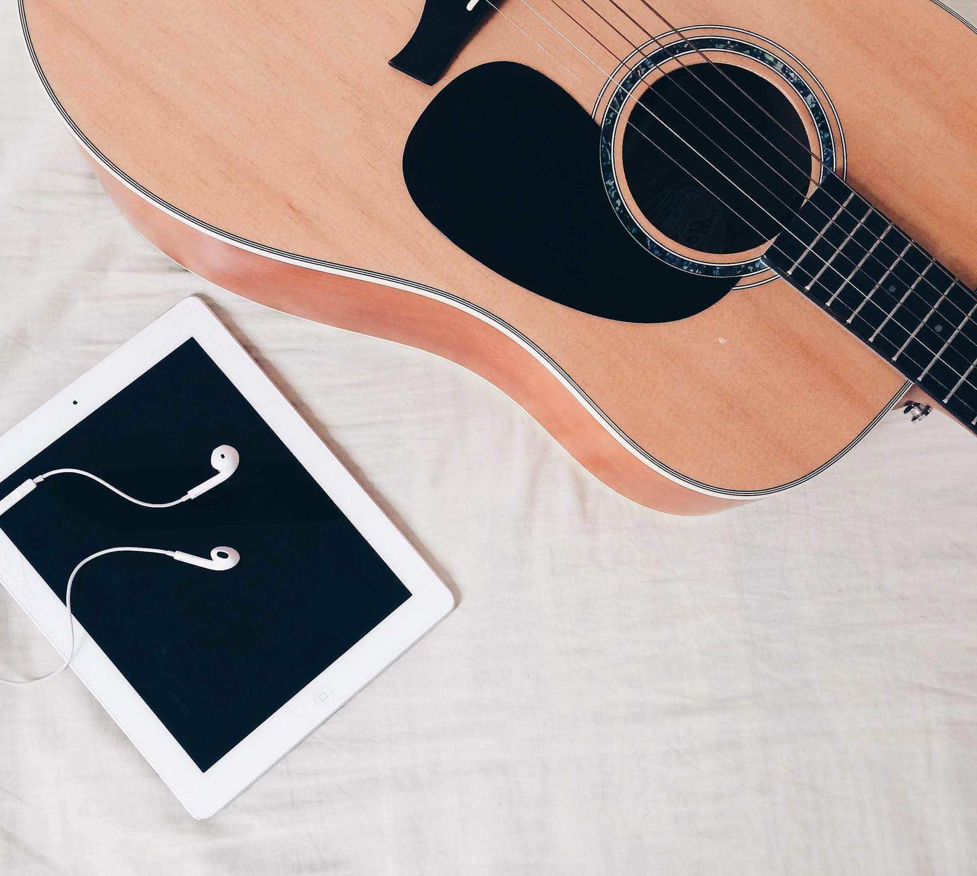 En lys gitar som ligger på et hvitt laken ved siden av en ipad