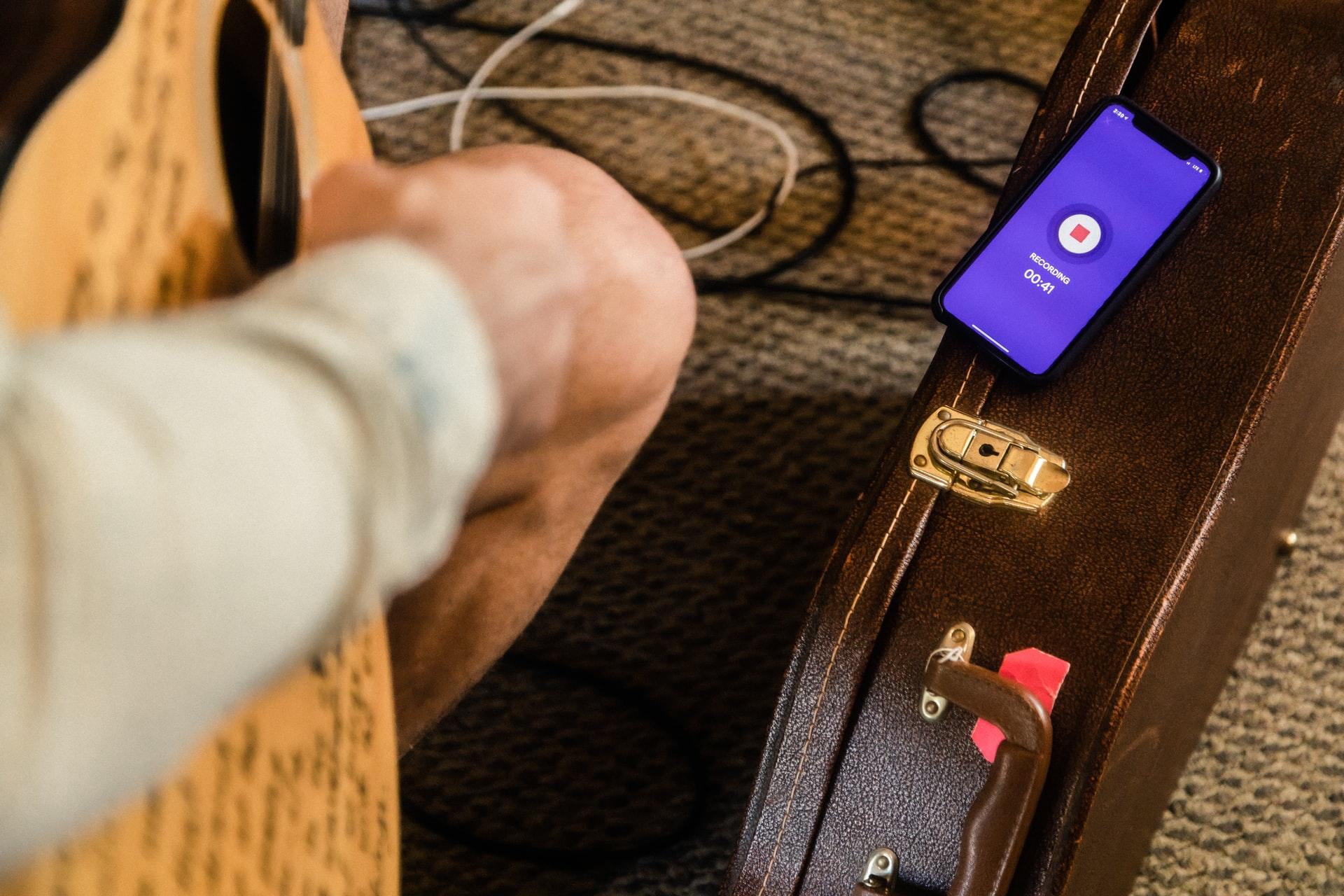 Nærbilde av en hånd som spiller på en gitar og en mobil som ligger oppå en gitarkasse
