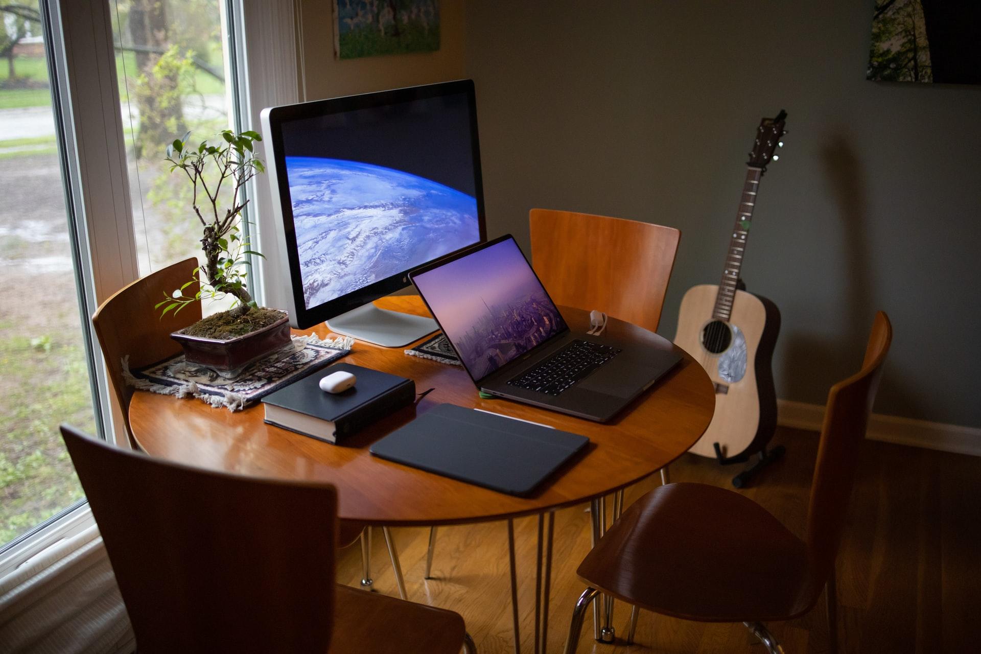 Et rom med et bord med fire stoler og en gitar rundt, og en laptop og pc-skjerm på