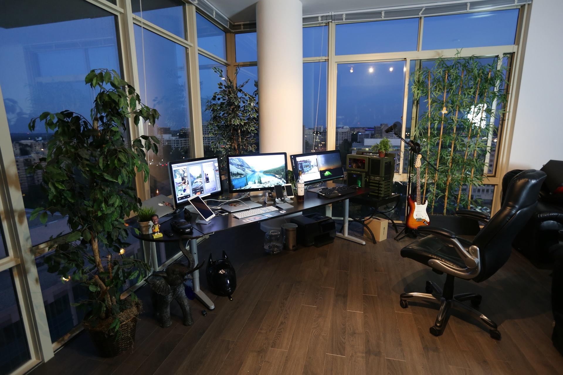 Et rom med store vinduer, planter og tre pc-skjermer på en pult som musikkutstyr