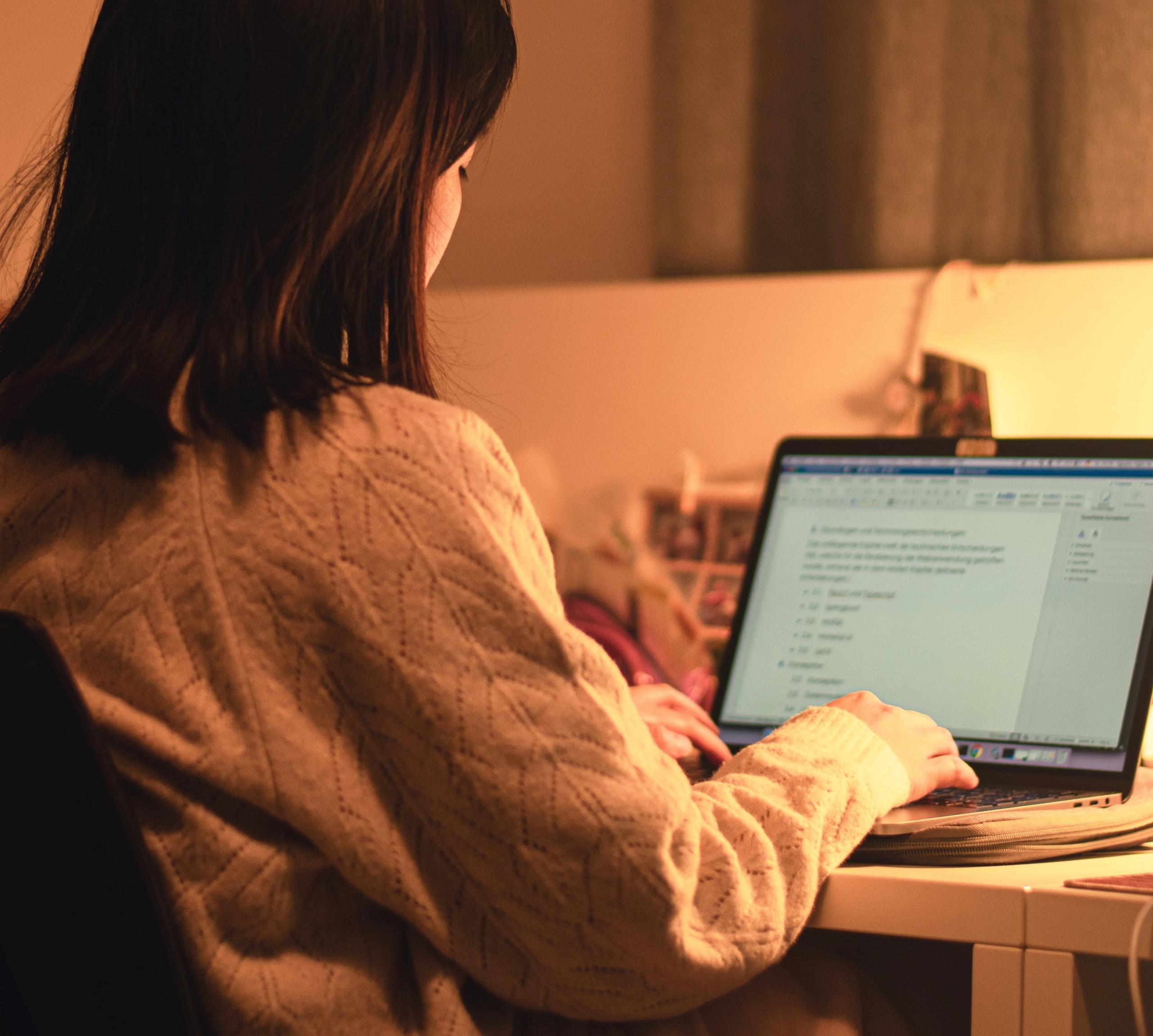 en ung kvinne sitter ved en laptop i varm innendørsbelysning og jobber